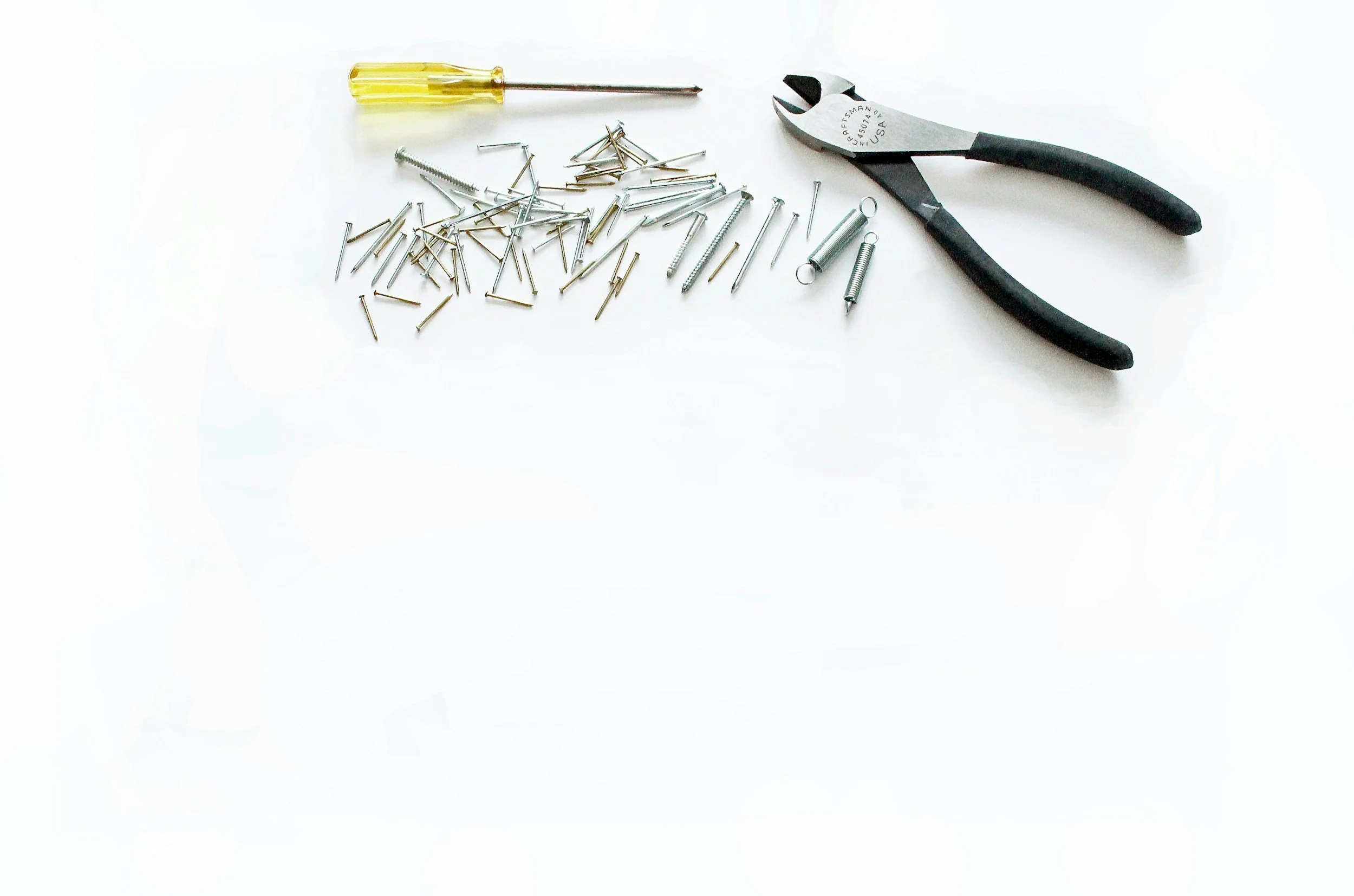 A white surface with a small yellow screwdriver, a pair of black-handled pliers, and various small metal nails and springs scattered around.