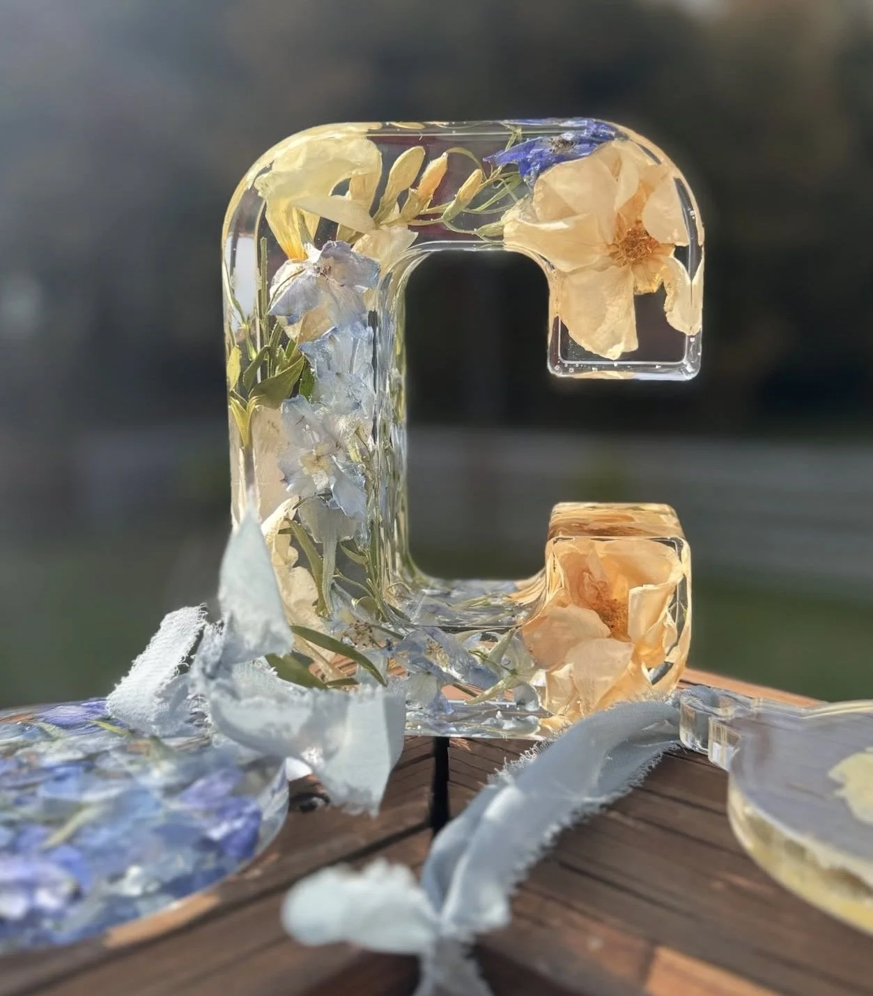 Letter C made of clear resin filled with dried flowers and leaves, placed on a wooden surface, with blurred outdoor background.