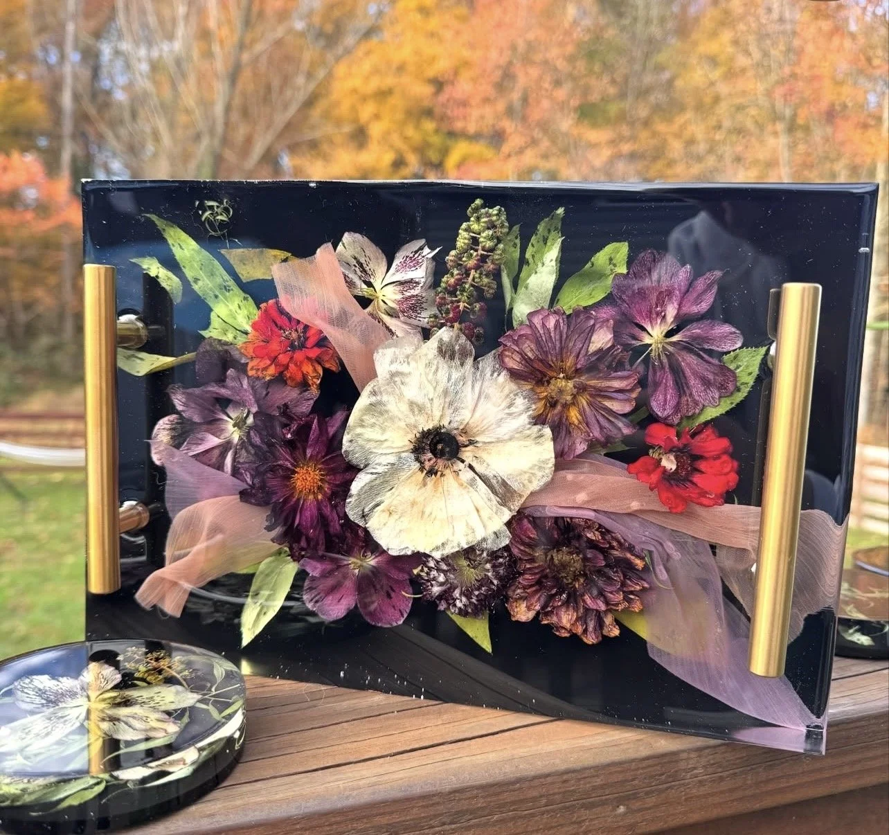 A transparent acrylic box with pressed flowers and leaves inside, adorned with pink ribbon and gold handles, placed on a wooden surface outdoors with trees and autumn foliage in the background.