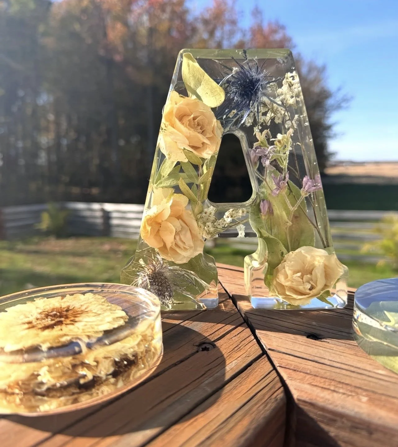 A decorative letter A made of transparent material filled with yellow roses, purple flowers, and greenery, placed on a wooden surface outdoors with trees and a fence in the background.