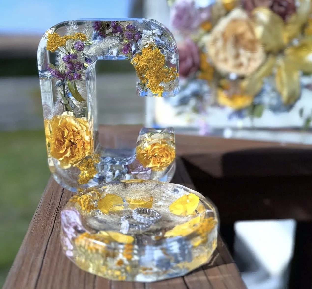 Decorative letter 'G' made of clear resin embedded with yellow, purple, and white flowers, set on a wooden surface with a floral arrangement in the background.