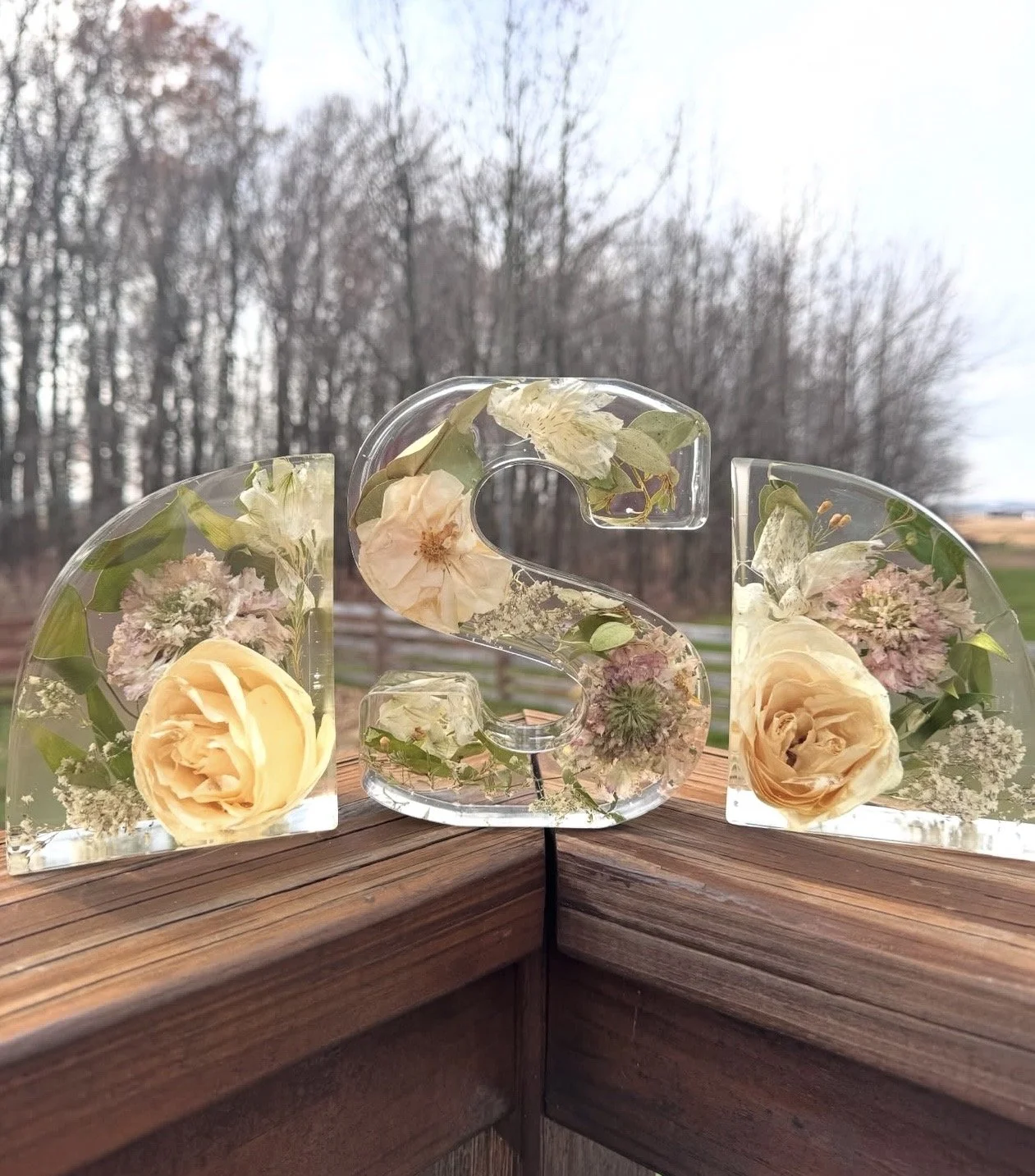 Decorative acrylic blocks with embedded flowers and foliage, arranged to spell 'ISA' on a wooden surface outdoors.
