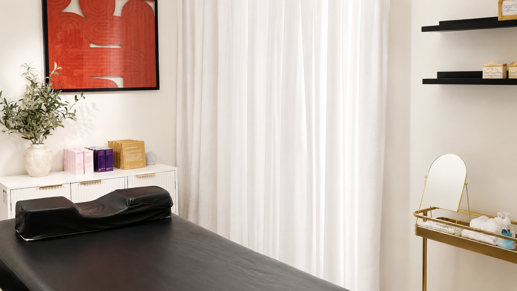 A wax studio with a black massage table, a white cabinet with pink, purple, and yellow packages, a potted plant, a large red abstract wall art, a white curtain, black floating shelves with boxes, and a golden tray with towels and bottles.