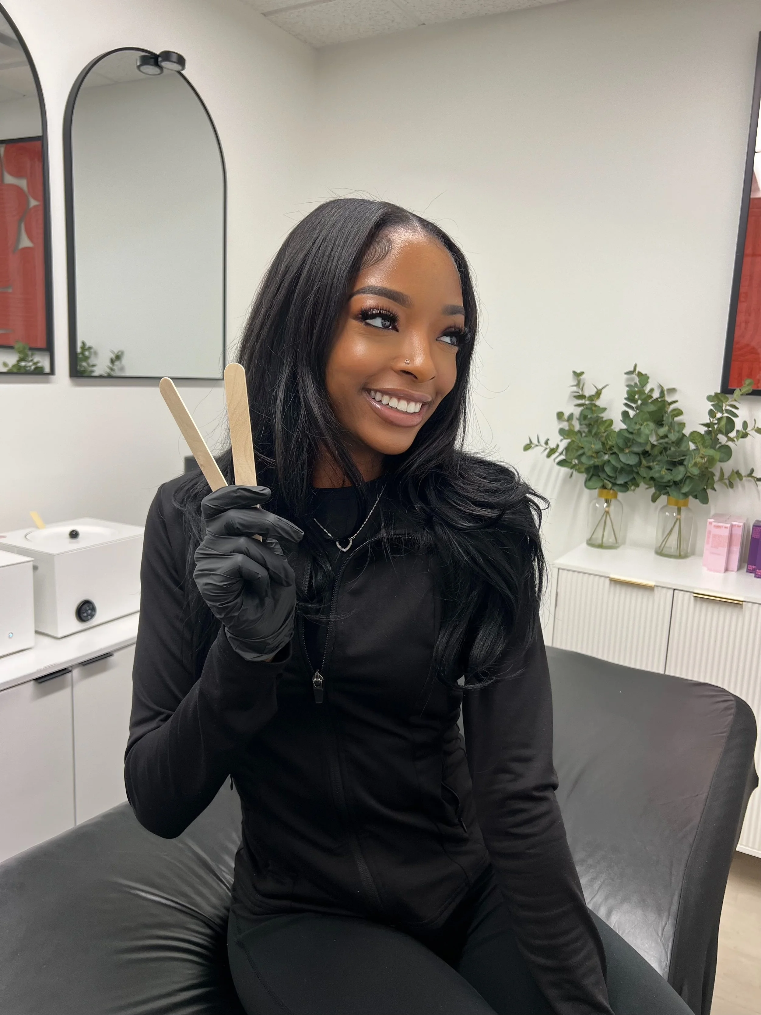 A woman with long black hair and a nose piercing smiling while holding two wooden popsicle sticks in her right hand, wearing a black jacket and gloves, sitting on a black leather couch in a room with white walls, mirrors, plants, and some decorative items.