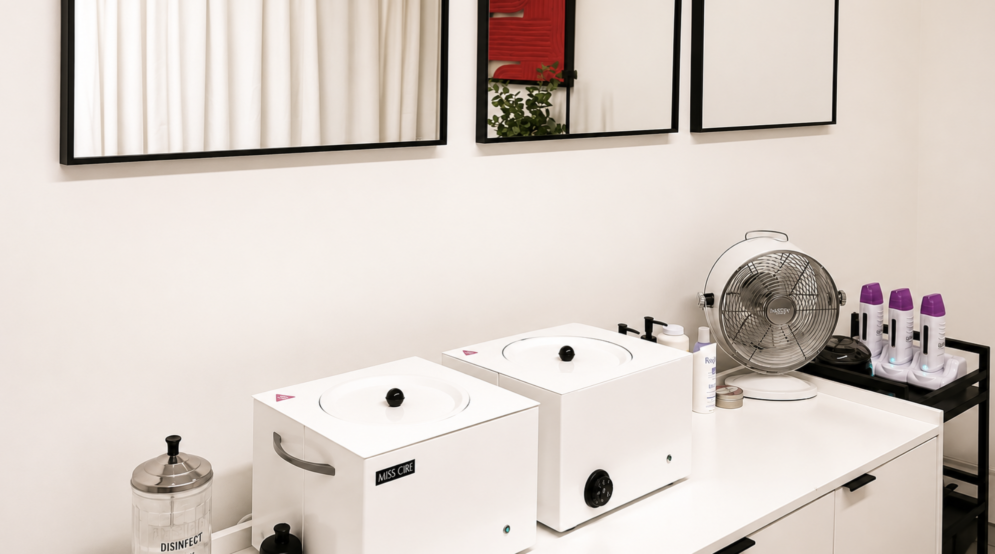 A white wax  station with two sterilizer units labeled 'MISS CIRE' on a white countertop, a fan, and multiple spray bottles. Black-framed mirrors and wall art are visible above the station, and the wall behind is plain white.