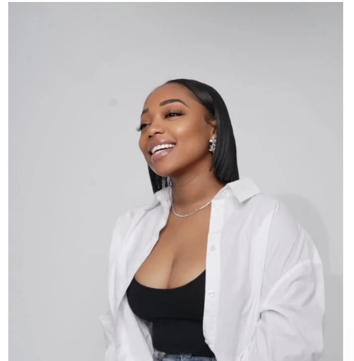 Smiling woman with black hair, wearing a white shirt over a black top, silver jewelry, posing against a plain background.