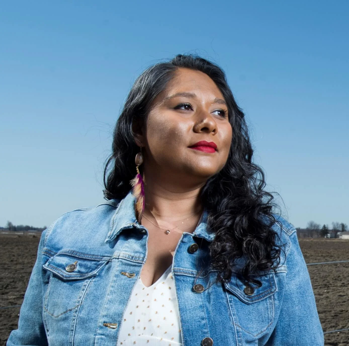 Roshan James wearing a denim jacket and a white top, standing outdoors against a clear blue sky with a rural landscape in the background.