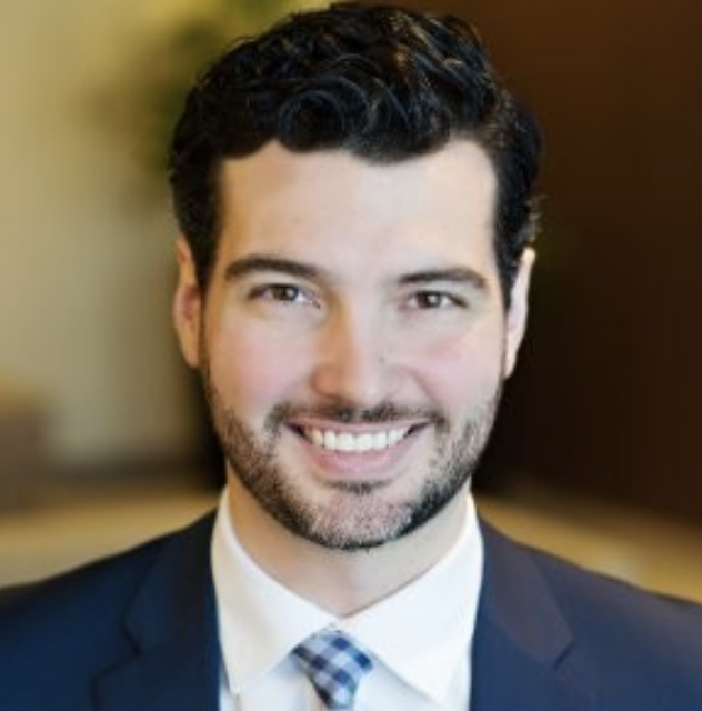 Peter Lowas in a dark suit, white shirt, and plaid tie smiling at the camera with a blurred background.