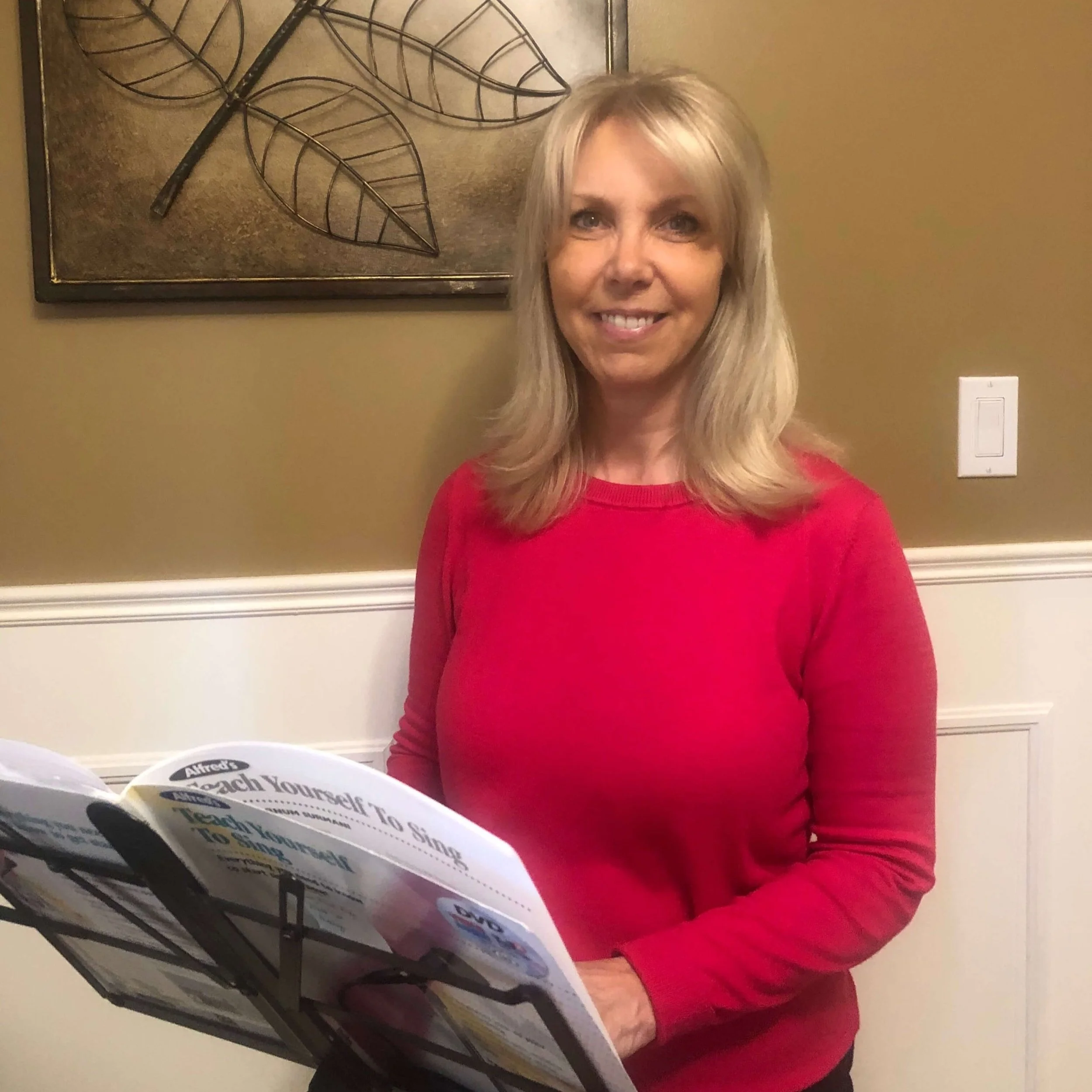 Tracie wearing a red long-sleeve shirt, standing indoors in front of a wall with artwork featuring leaves, holding a book or pamphlet titled 'Teach Yourself To Sing'