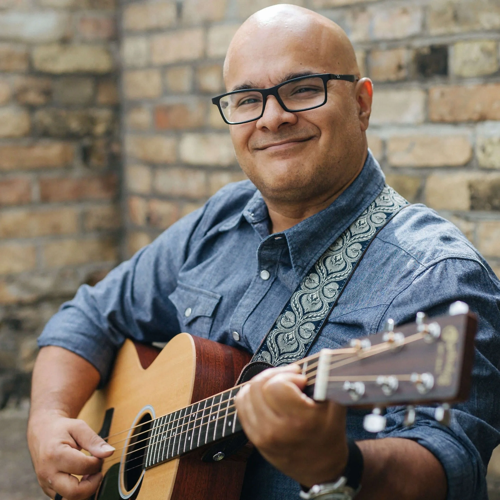 Martin playing an acoustic guitar, smiling, against a brick wall background.