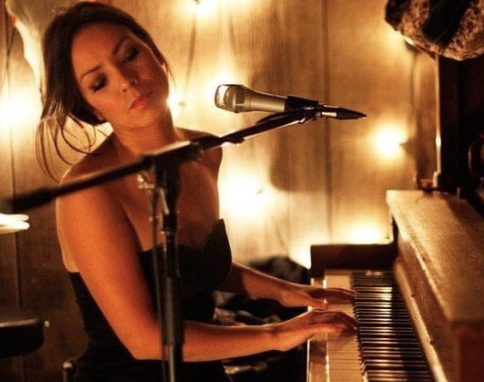 Emm Gryner in a black dress playing a piano and singing into a microphone, in a dimly lit room with warm lighting.