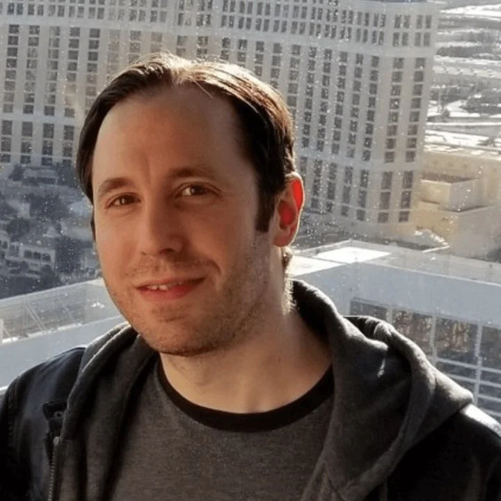 Eric, smiling, wearing a black jacket and a dark T-shirt, standing in front of a cityscape with tall buildings and glass windows.