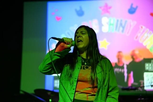Christine singing into a microphone on a stage with a colorful background that says 'Shiny Things' and has various decorative icons.