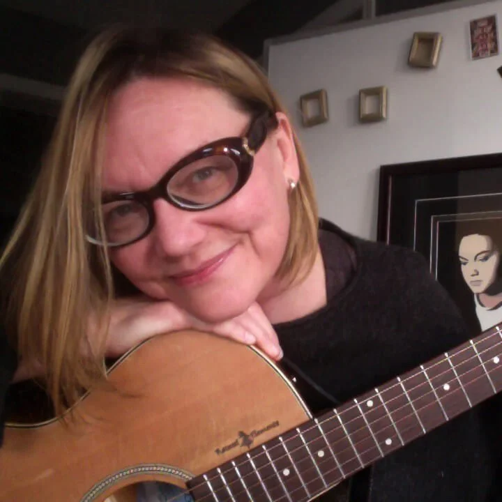 Cheryl smiles while holding an acoustic guitar, leaning her head on it in a cozy room with framed art on the wall.