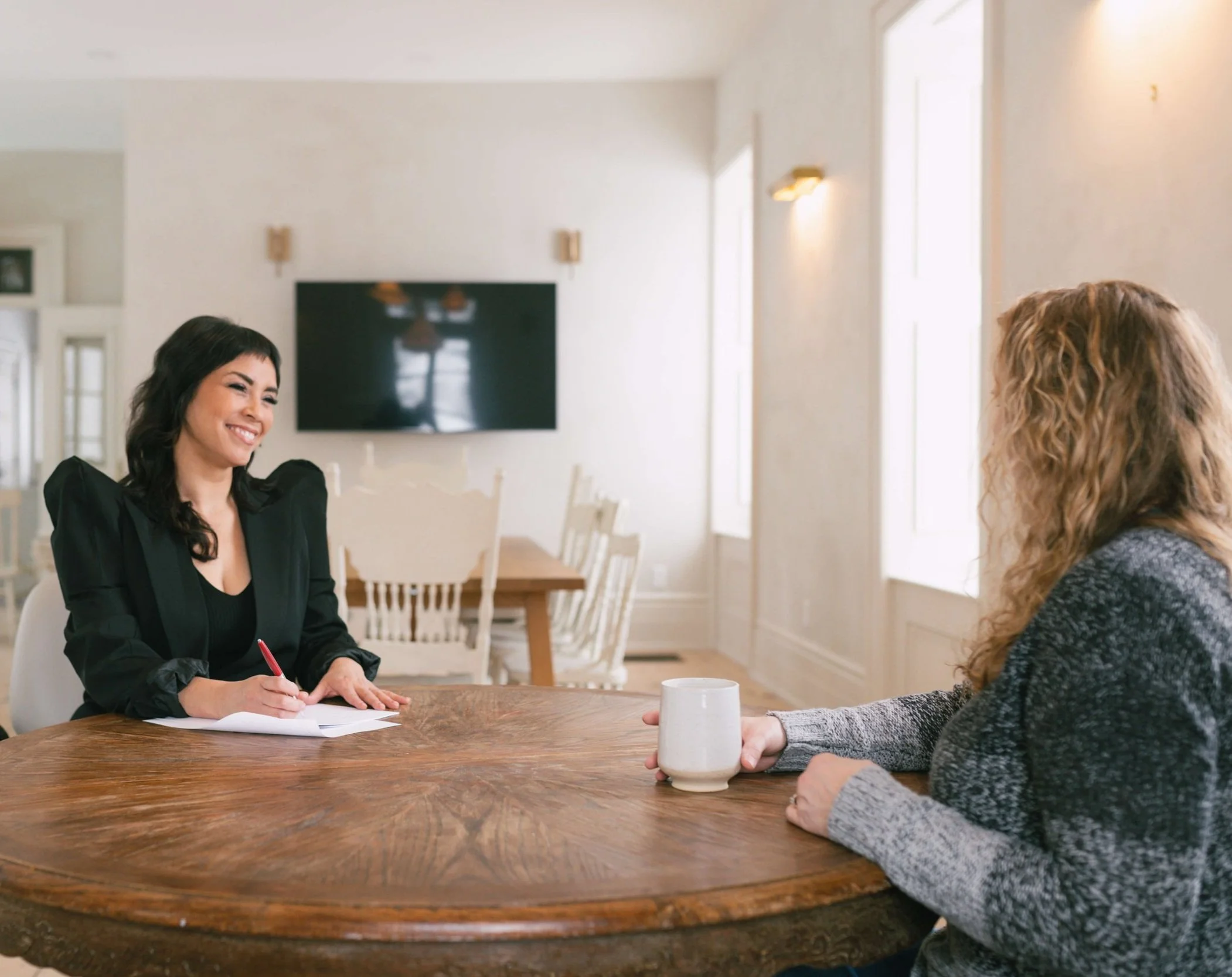 Emm Gryner having a conversation at a wooden dining table in a well-lit room, one with dark hair, wearing a black blazer, smiling, and taking notes with a student with wavy hair, wearing a gray sweater, holding a white mug.