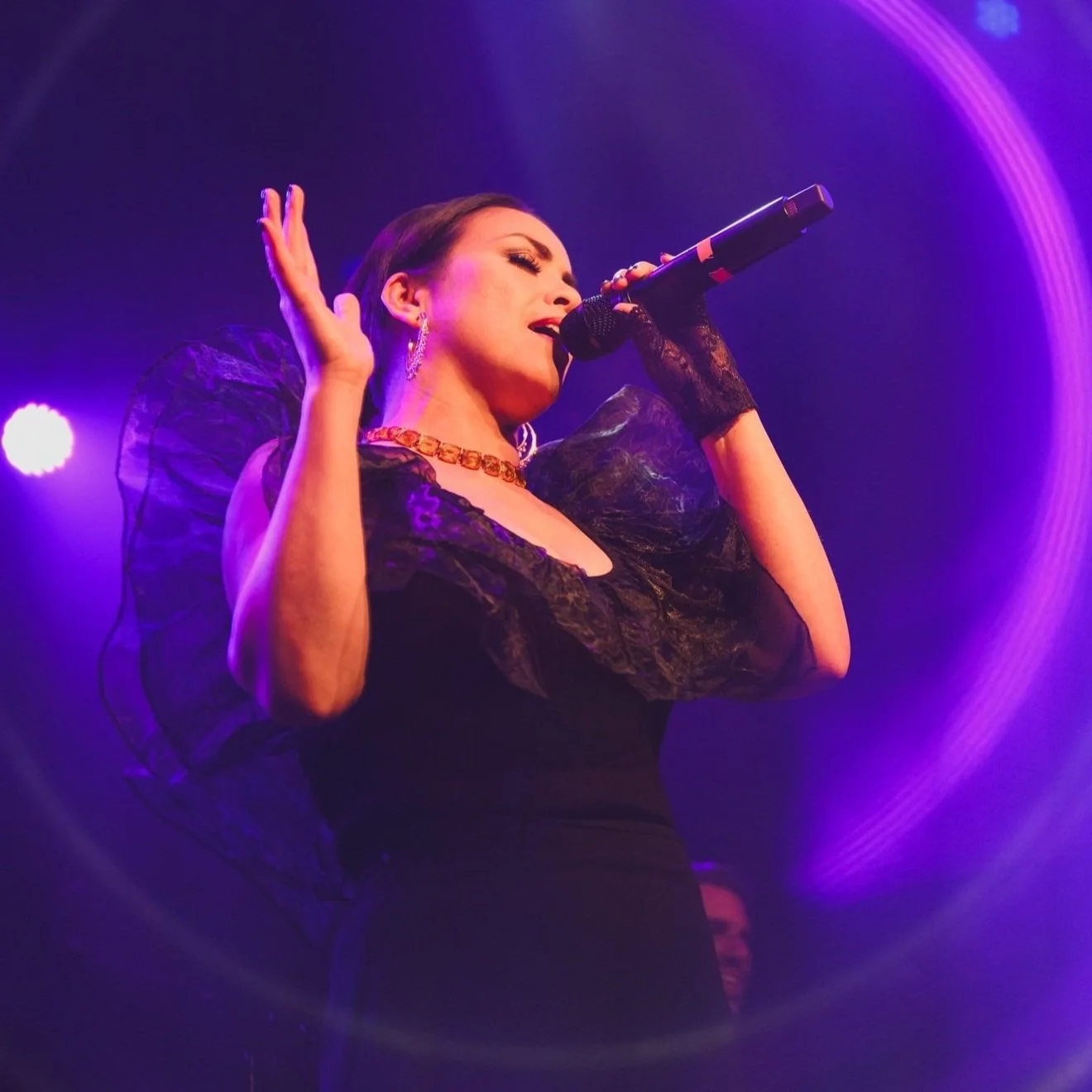 Emm Gryner into a microphone on stage, bathed in purple and blue stage lights, wearing black lace gloves, a black dress with sheer puffed sleeves, and gold jewelry.