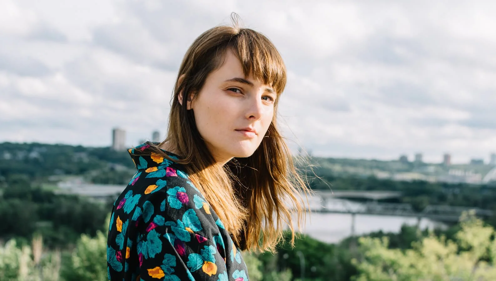 Rachel, Pro Writer & Recording Artist looking at the camera, wearing a colorful floral jacket, outdoors with a blurred cityscape and cloudy sky in the background.