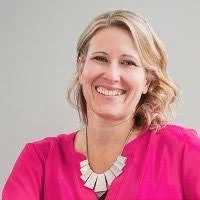 Emm Gryner wearing a black blazer with exaggerated shoulder pads, looking confidently at the camera against a red circular background.