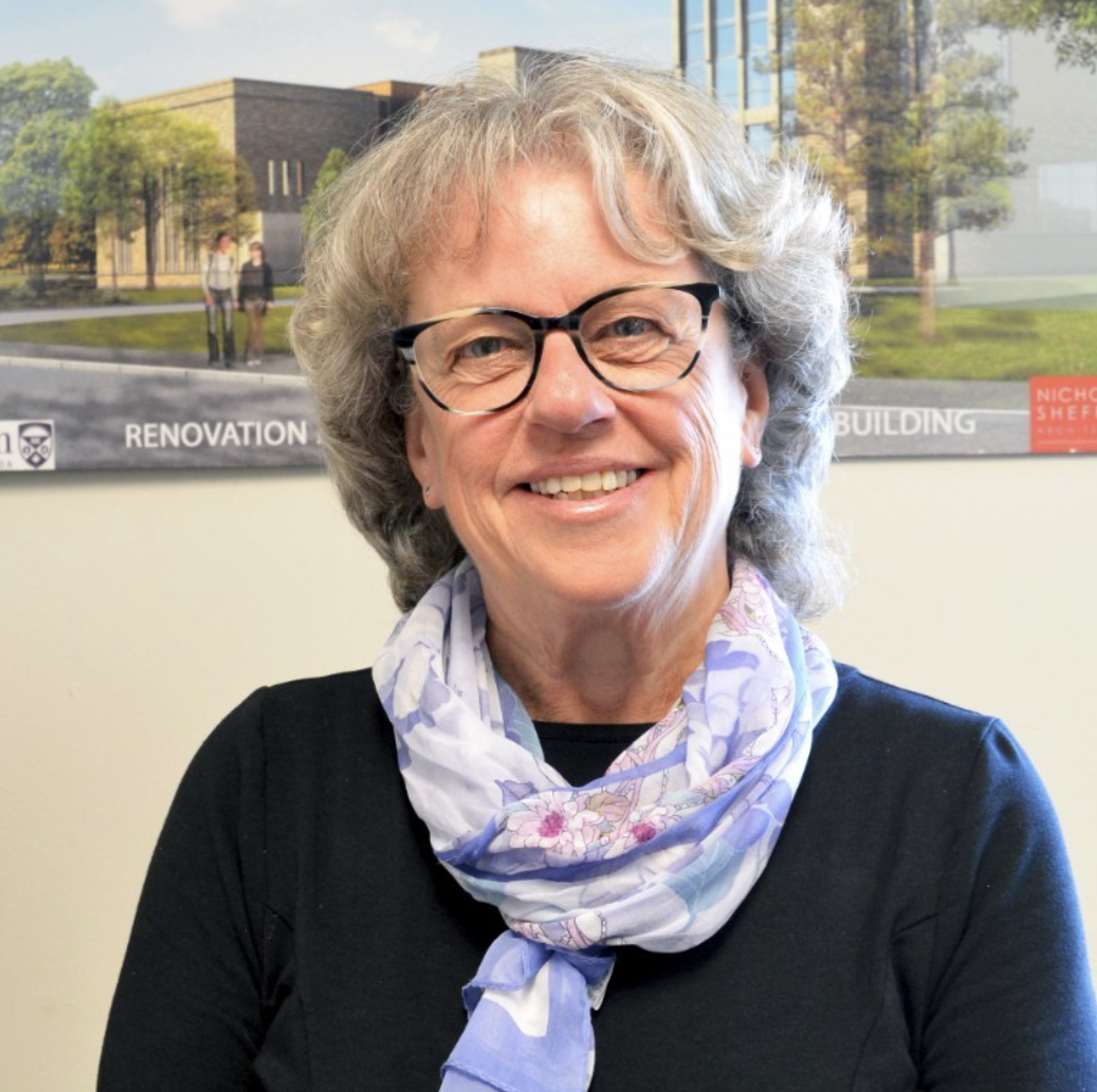Betty Anne Younker, wearing a black top and a light purple floral scarf, standing indoors with a campus or academic building visible on the wall behind her.