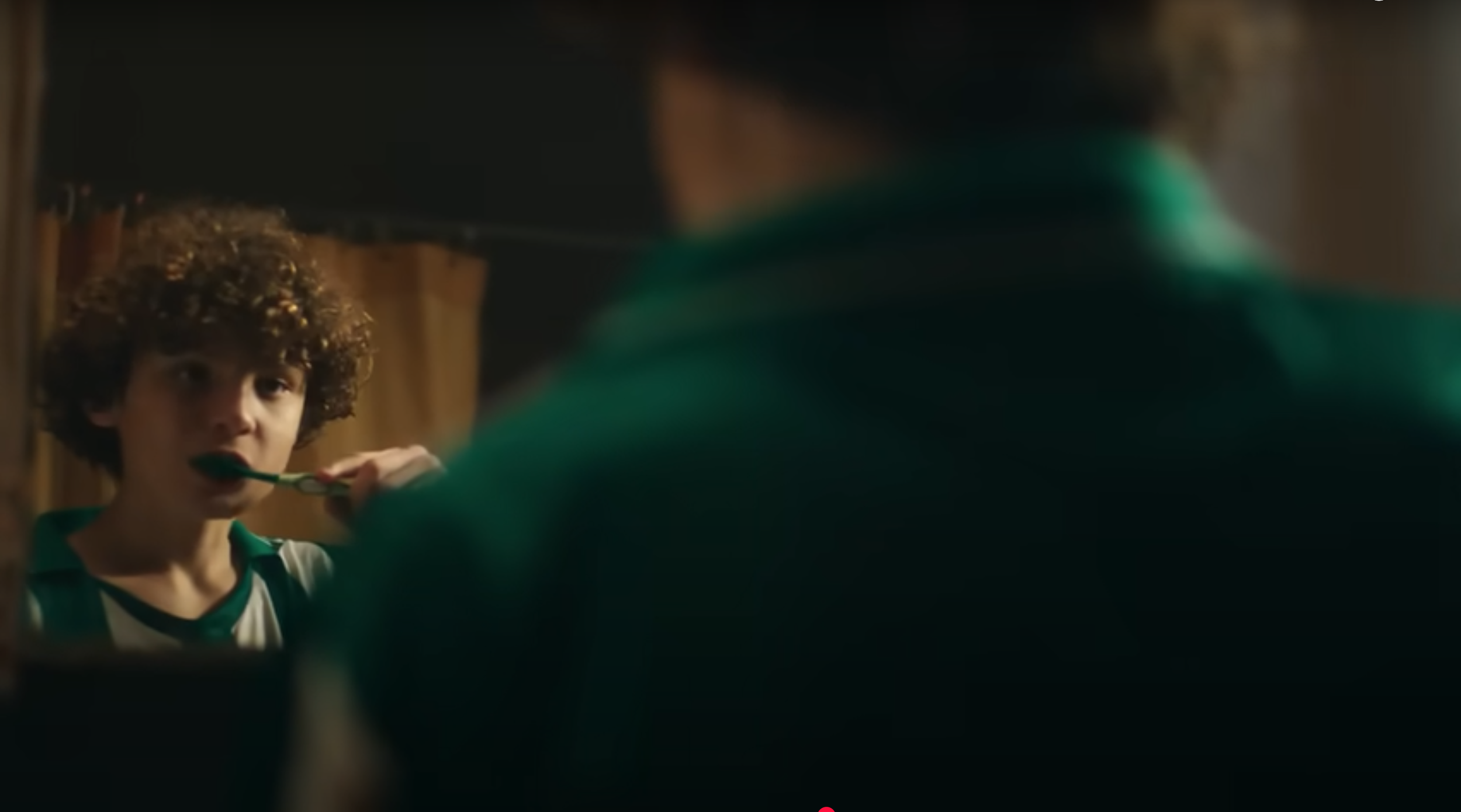 Un niño con cabello rizado mira hacia el espejo mientras se cepilla los dientes, reflejado en un espejo en un entorno que parece un baño o habitación