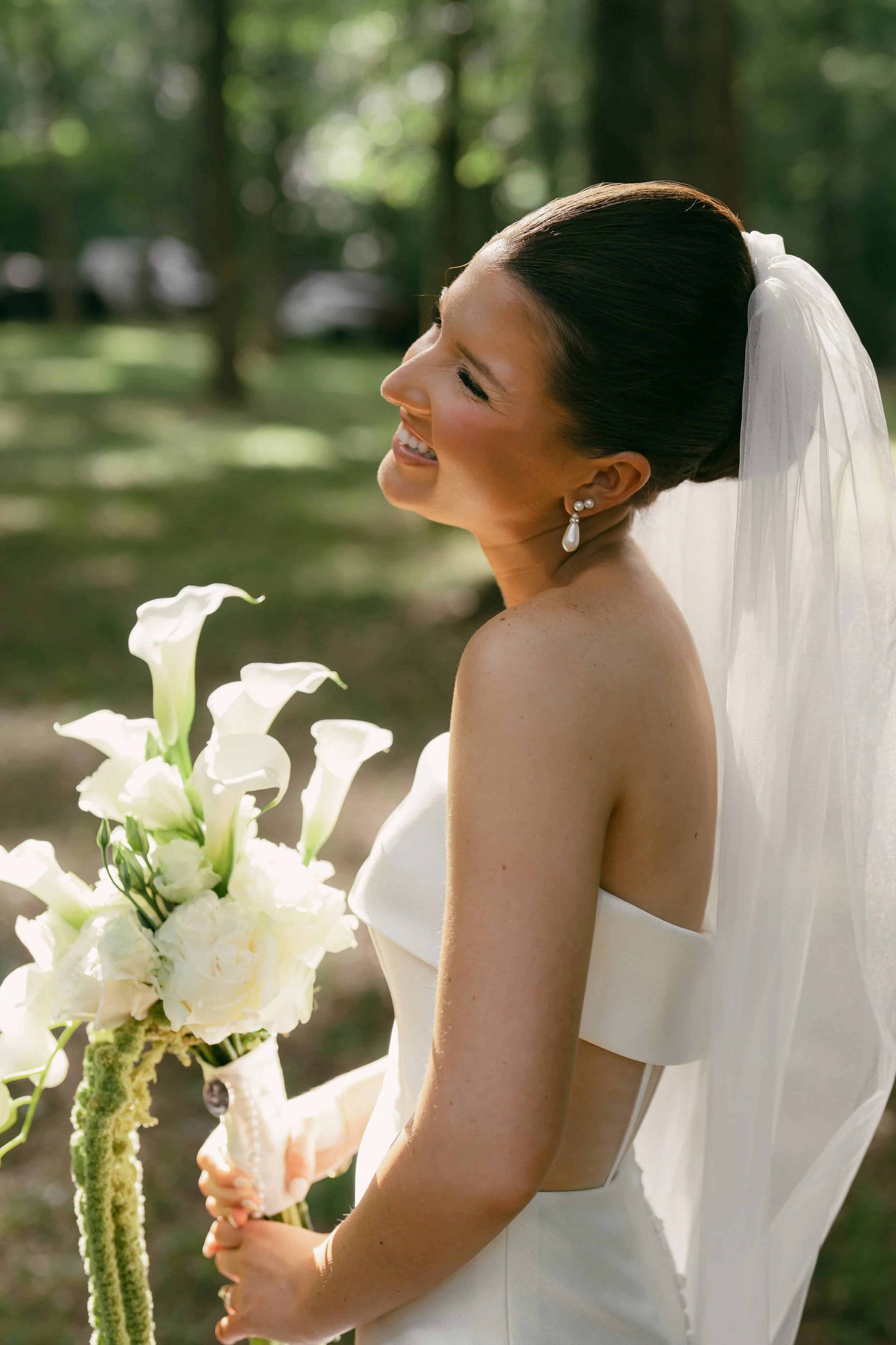 A bride smiling outdoors, holding a bouquet of white calla lilies, wearing a strapless white dress and pearl earrings, with a veil and her dark hair styled in an elegant updo.