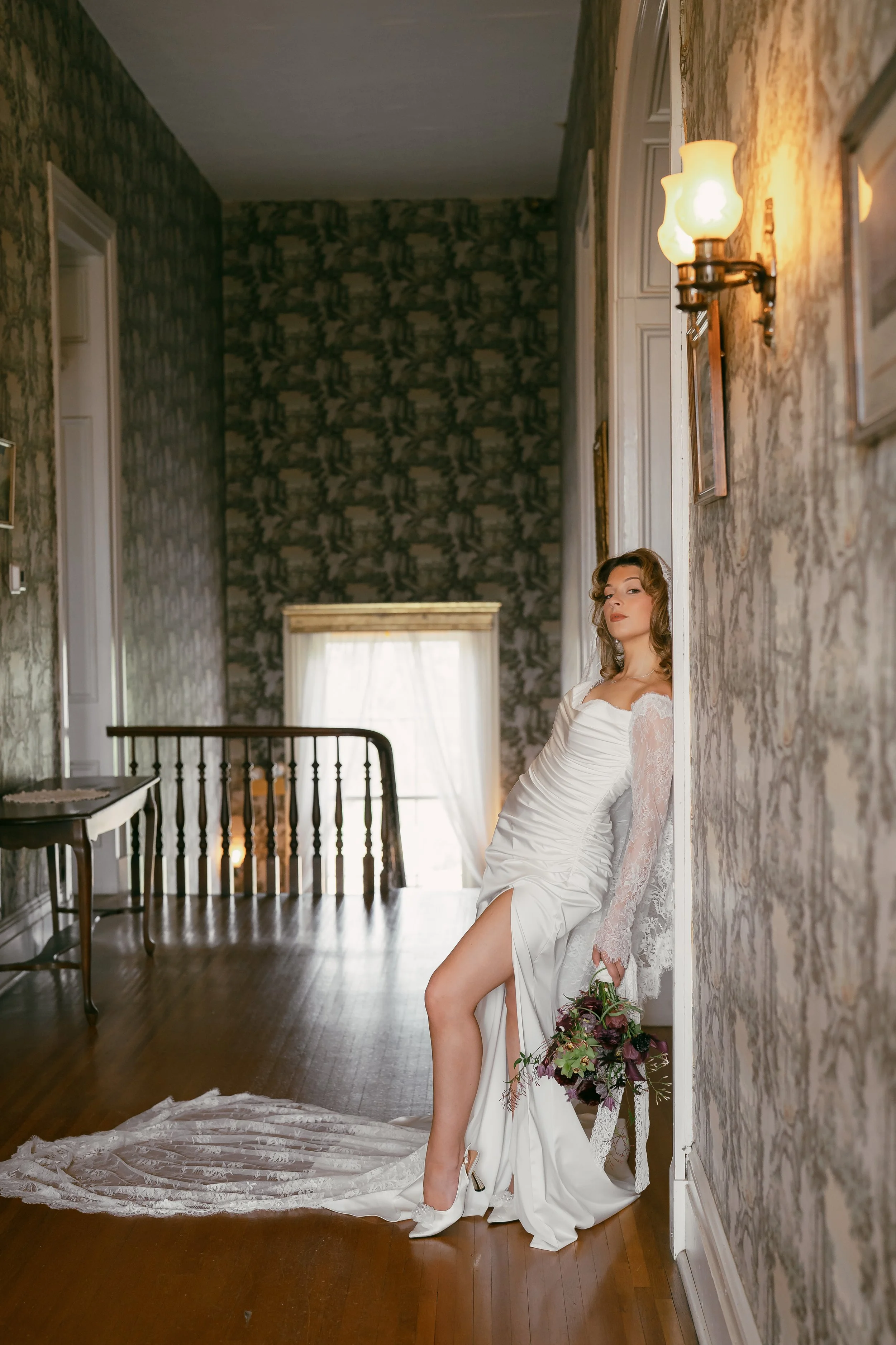 Woman in a wedding dress with a floor length, cathedral veil leaning against a wall in a vintage-style room, holding a bouquet of flowers, with her dress extending onto the wooden floor.