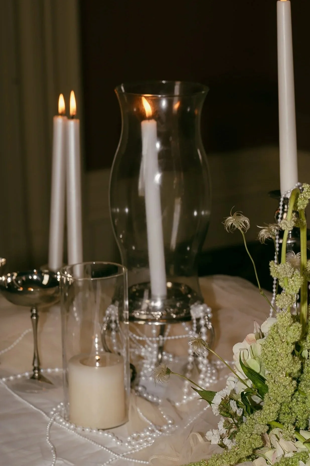 Decorative table centerpiece with candles, a glass vase, flowers, and pearl garlands for an elegant wedding reception.