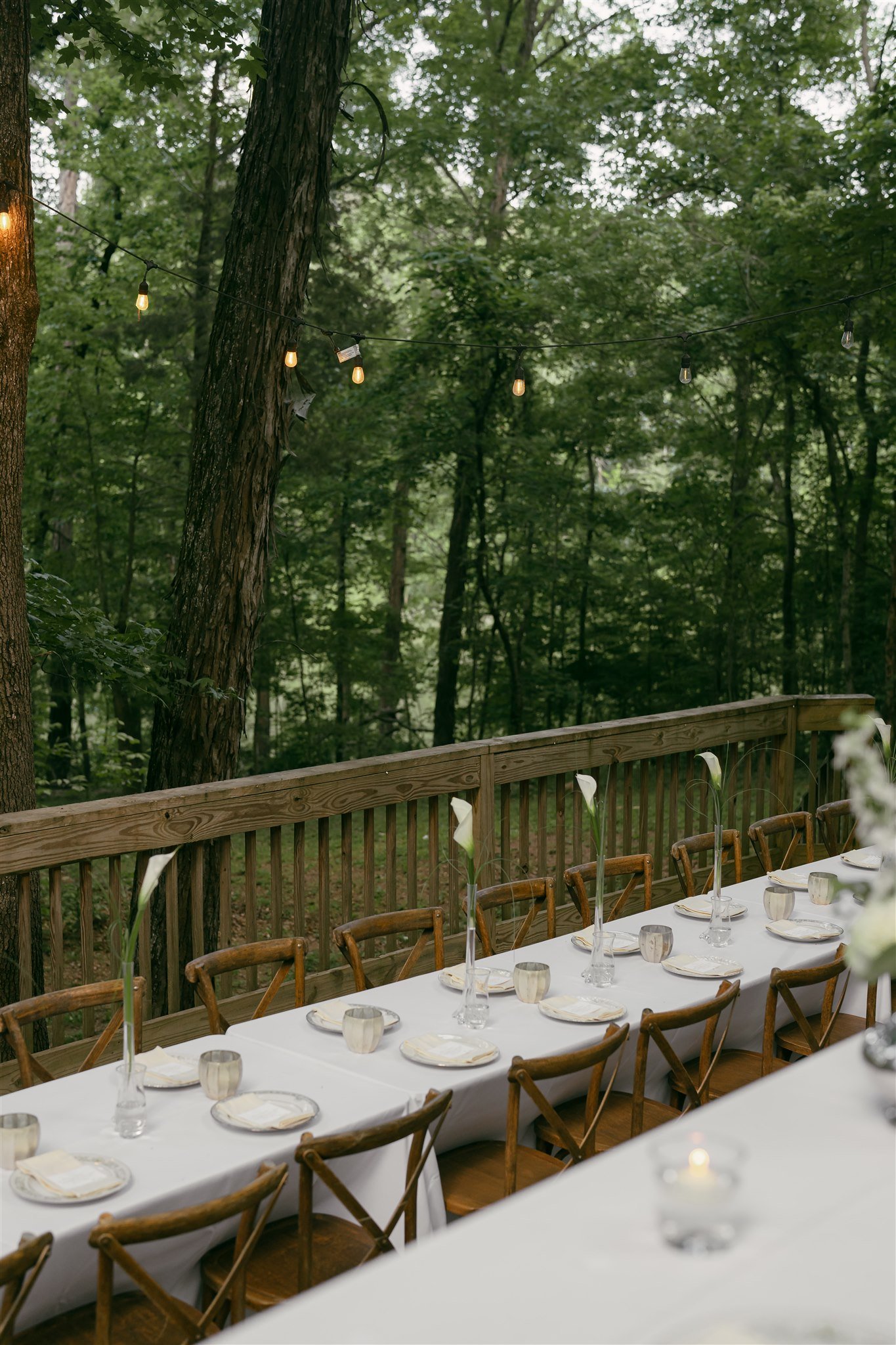 A long outdoor dining table set for a wedding reception, with  china plates, glasses, and small vases with calla lily flowers, surrounded by wooden chairs, with a forested background and string lights hanging overhead.