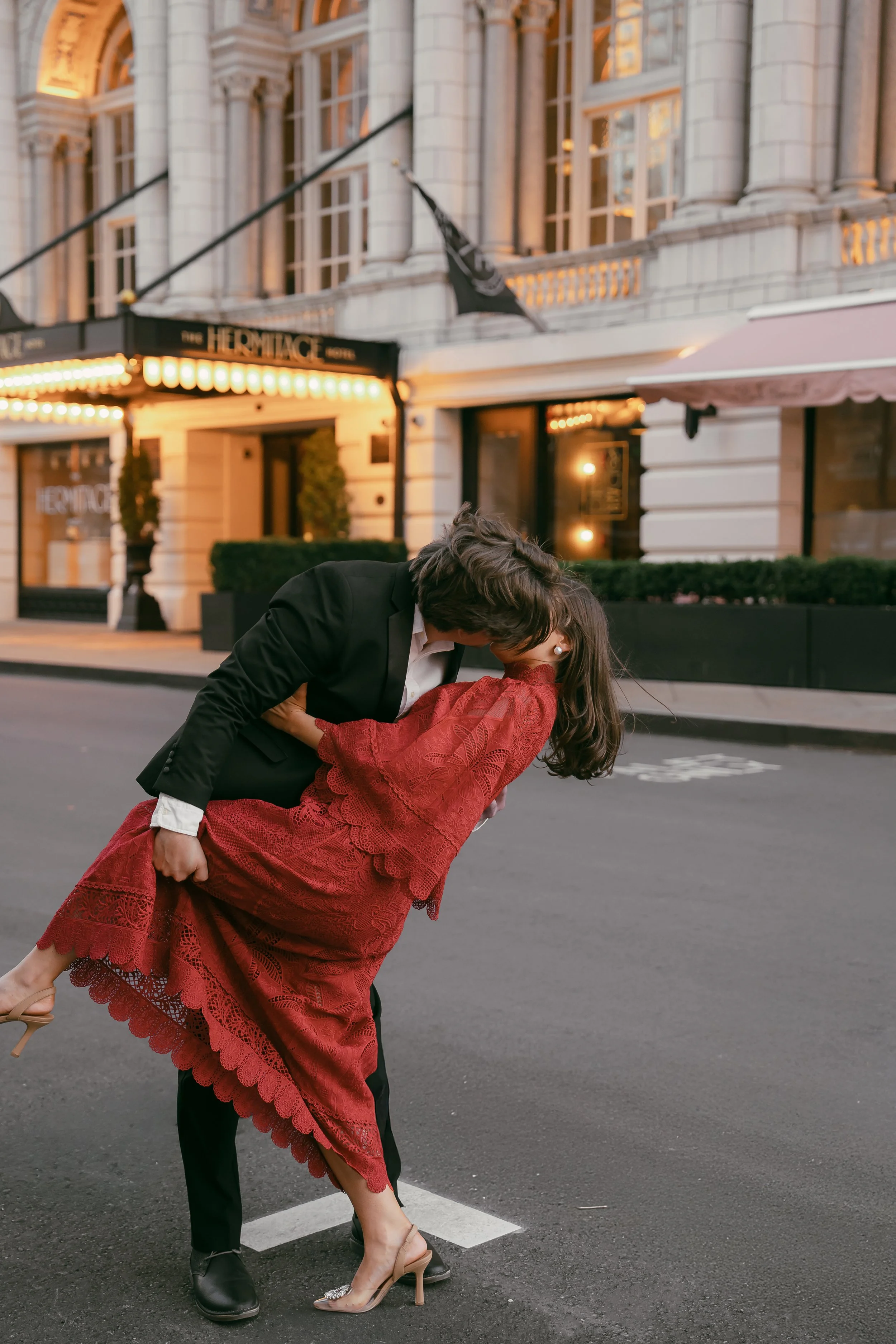 A man in a black suit dips a woman in a red dress on an empty city street at dusk, near The Hermitage Hotel with glowing lights in Nashville, Tennessee.