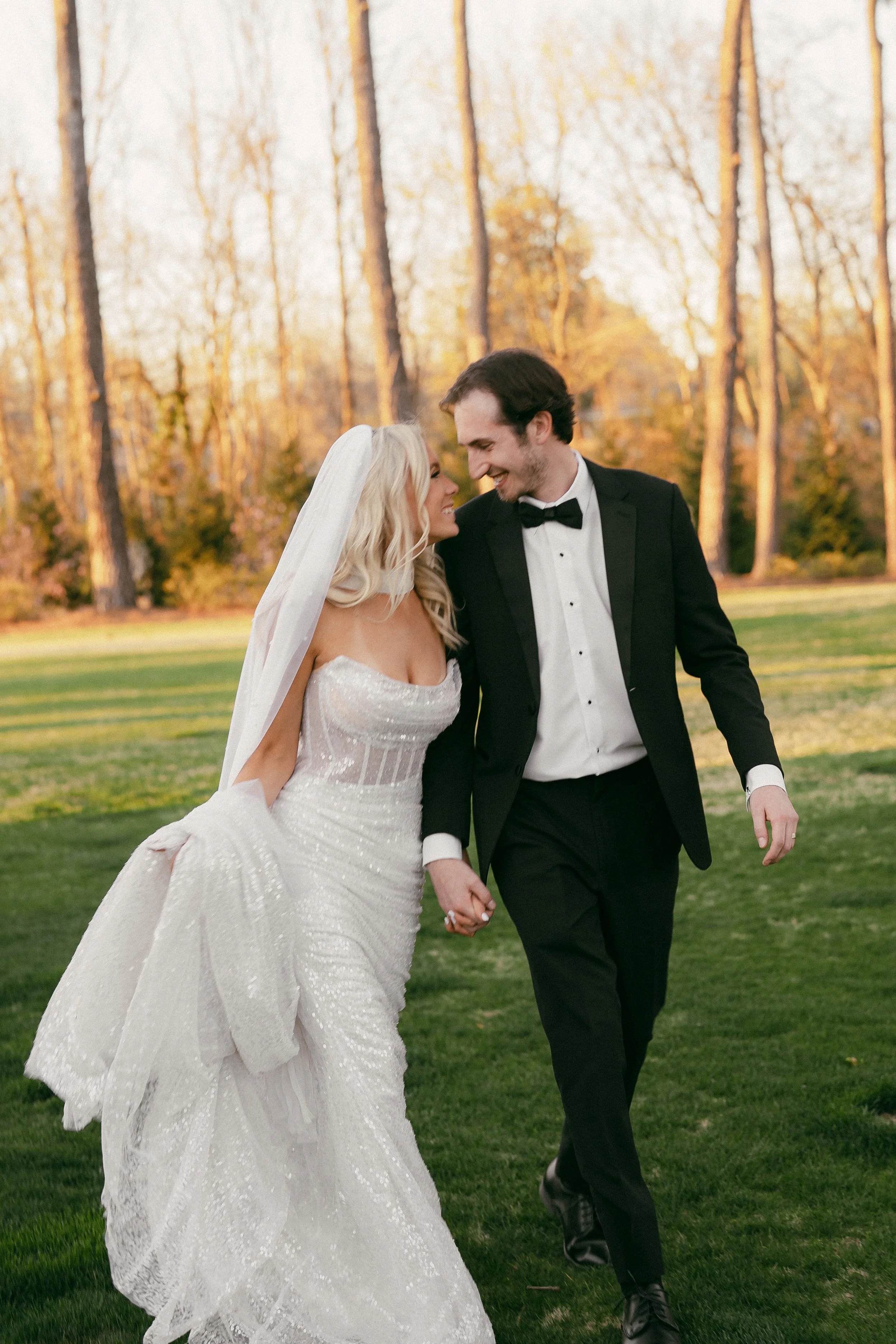 A newlywed couple holding hands and smiling, walking on a grassy area outdoors with trees in the background, during sunset at The Lumpkin House Venue in Albertville, Alabama.