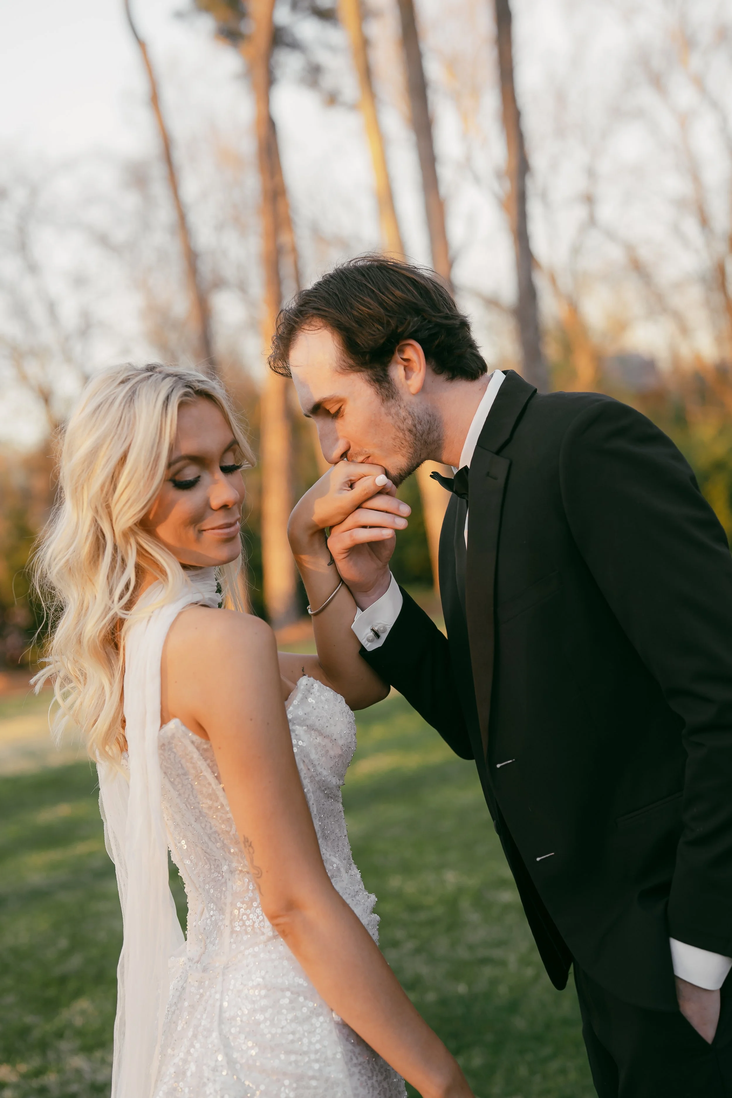 A bride and groom in wedding attire outdoors, with the groom kissing the bride's hand at The Lumpkin House Venue in Albertville, Alabama.