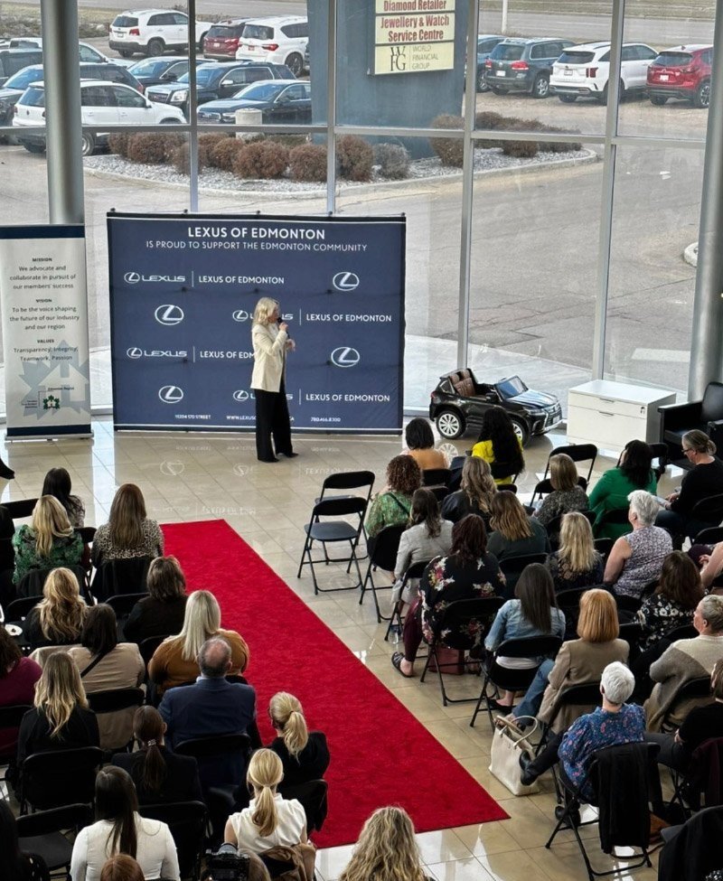 A speaker addressing a seated audience in front of a Lexus of Edmonton backdrop with a red carpet in a showroom.