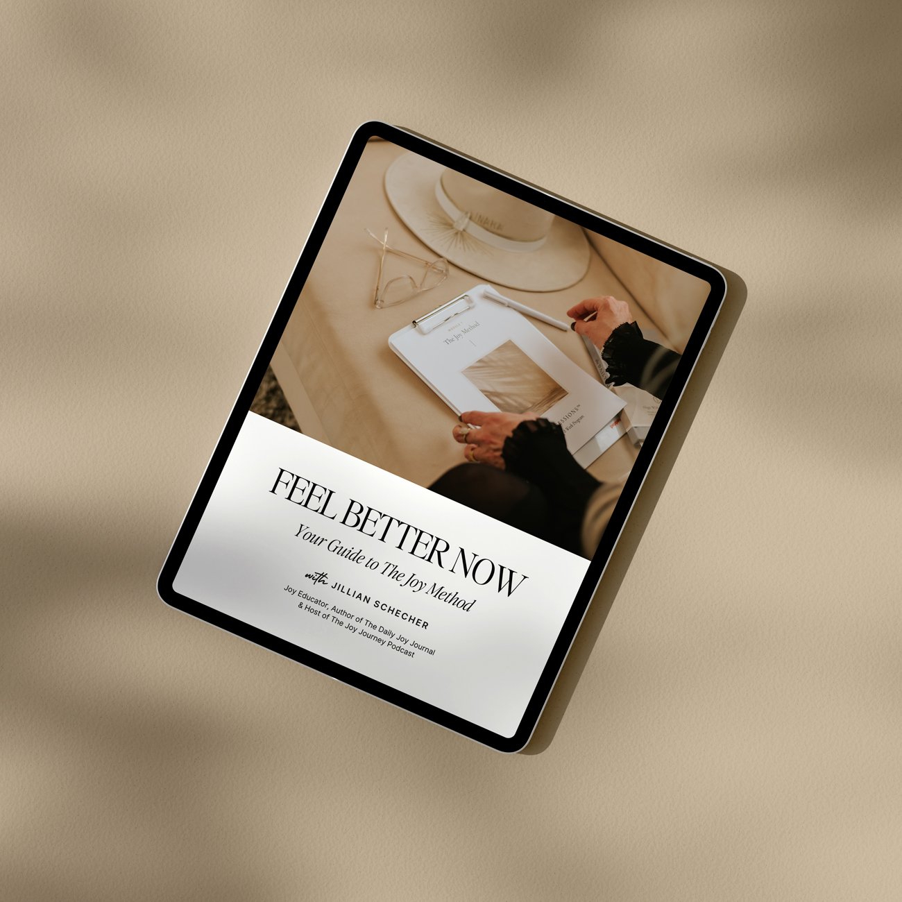 Tablet displaying "Feel Better Now: Your Guide to The Joy Method" by Jillian Scherer, featuring a person writing on a clipboard with a hat and glasses nearby on a beige table.