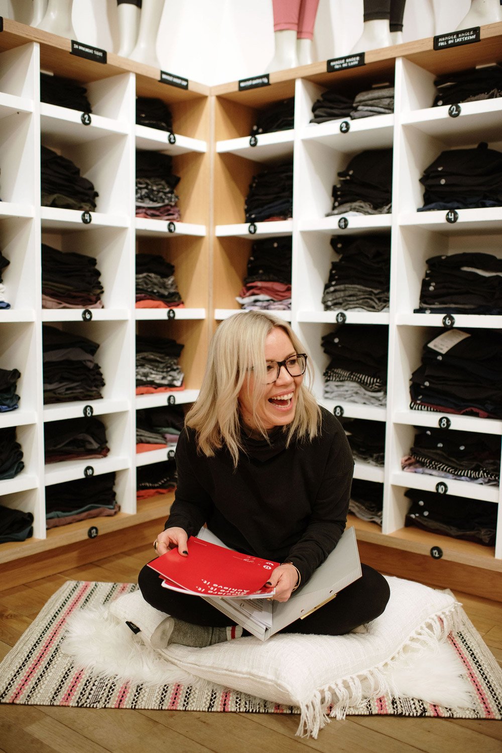 Woman sitting on floor in front of shelves with folded clothes, smiling and holding papers.
