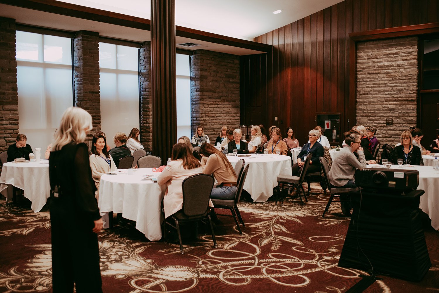 People attending a seminar in a conference room with round tables and brick walls.