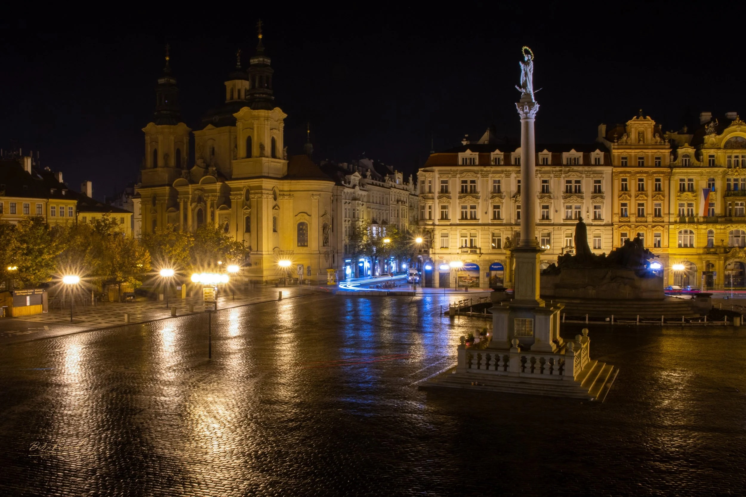 Night view of Old Town square in Prague with a monument featuring a tall column topped by a statue, surrounded by historic buildings and lit streetlights reflecting on wet cobblestone pavement.