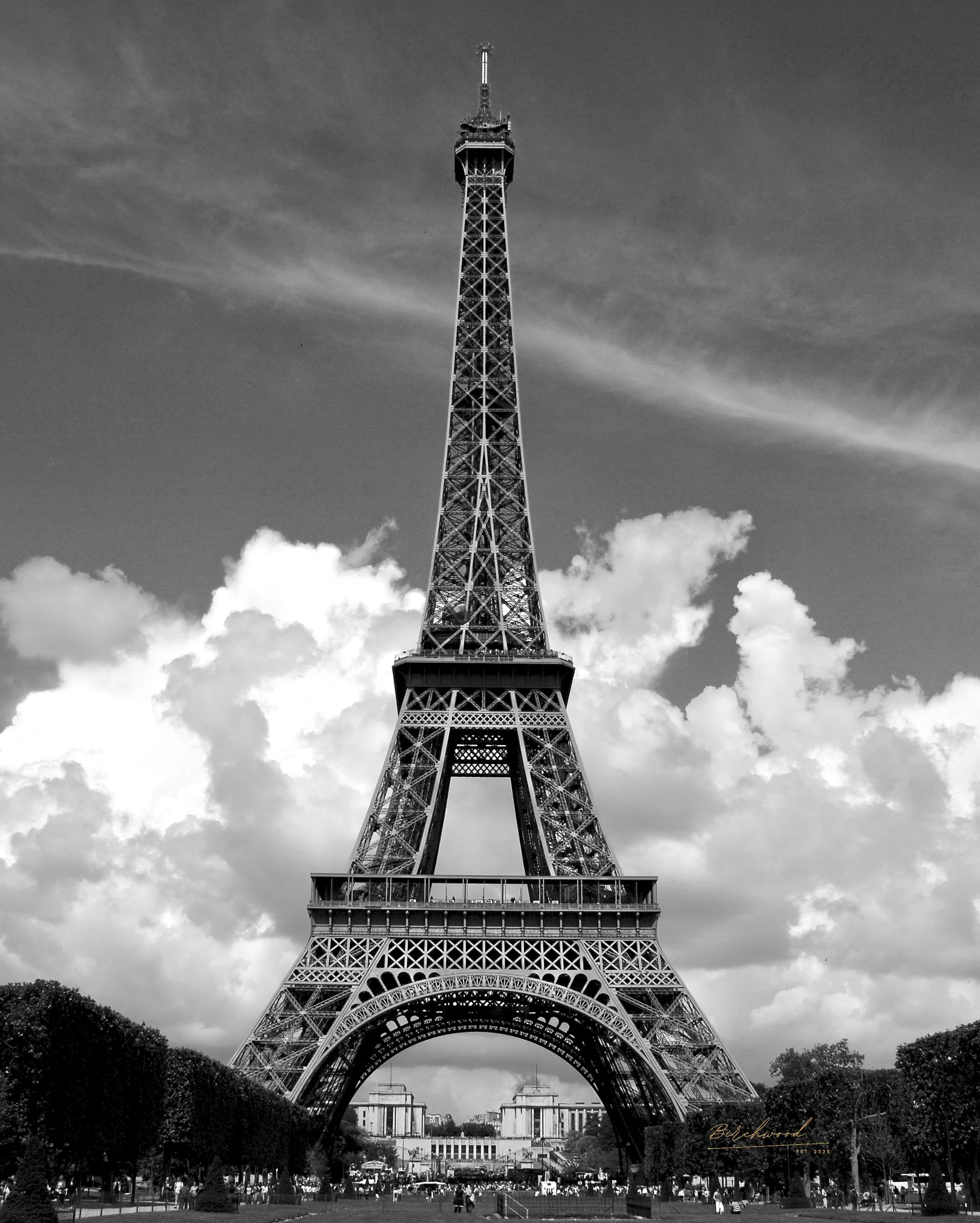 Black and white photo of the Eiffel Tower in Paris, France, with a cloudy sky overhead and people gathered at its base.