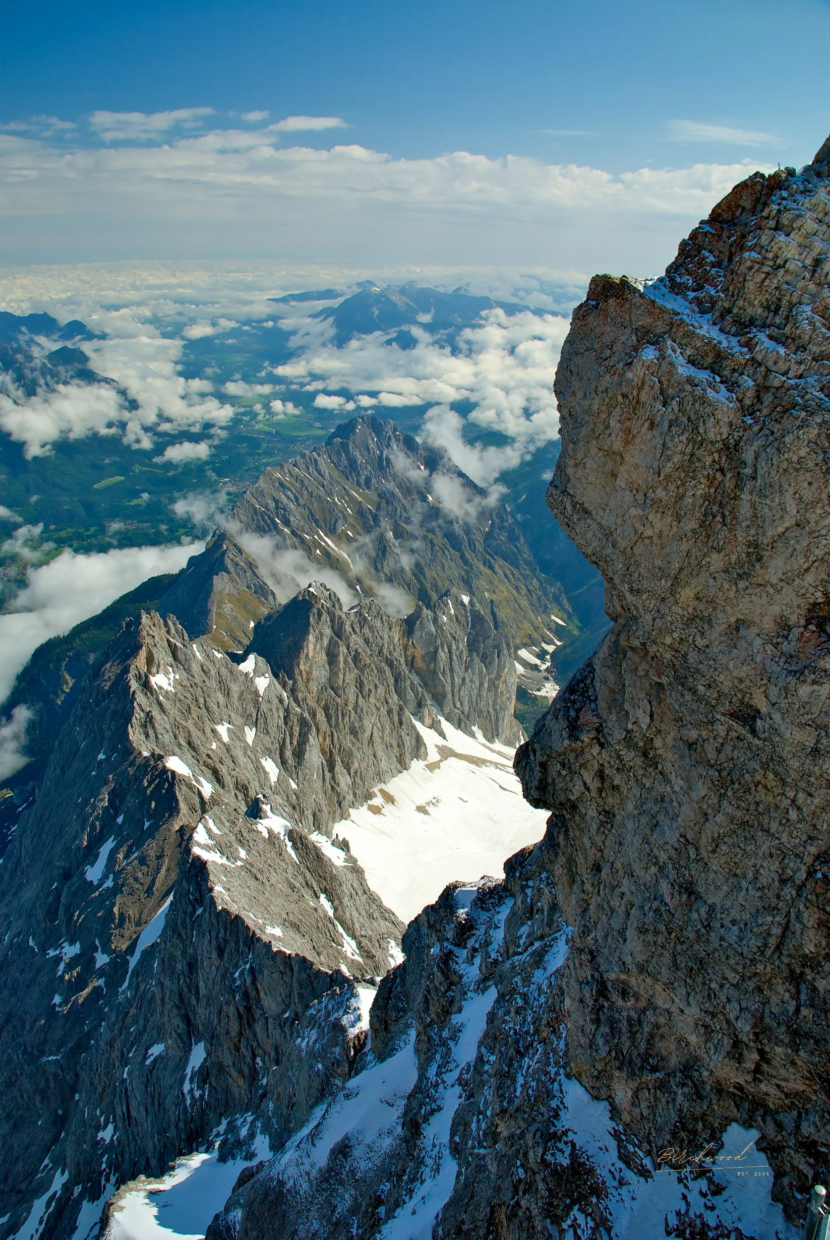 View from the Zugspitz in the German Alps with snow patches, clouds, and distant peaks under a blue sky.