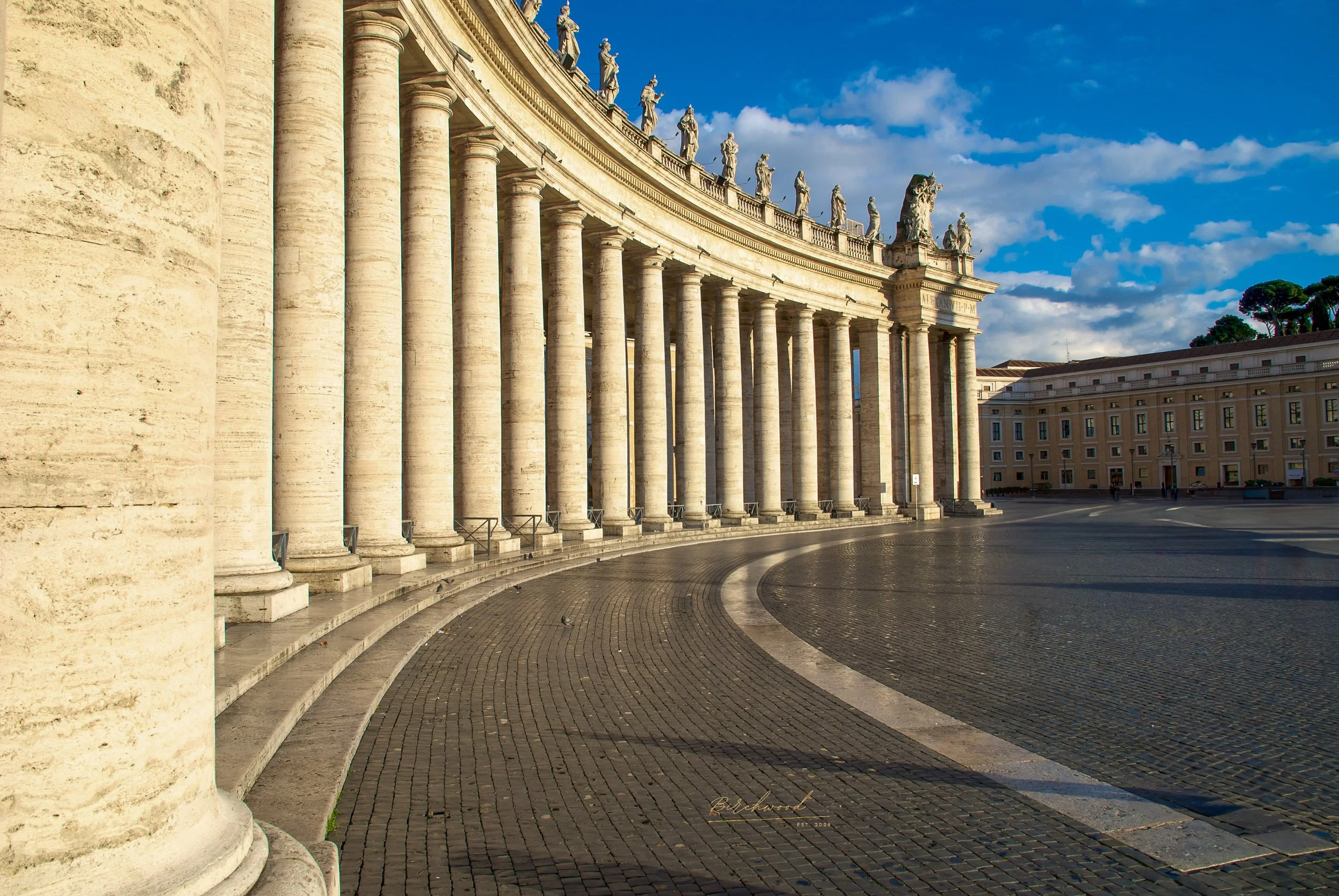 Photograph of a colonnade in the Vatican with tall, white stone columns and statues on top, curved architecture, under a partly cloudy sky, with a paved open area in front.