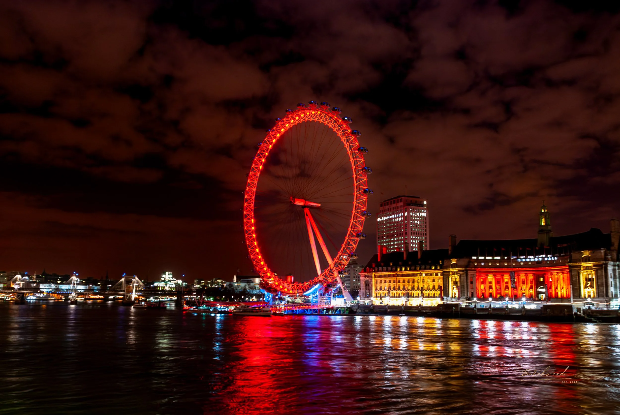 Night view of the London Eye illuminated in red, reflecting on the river with buildings in the background under a cloudy sky.
