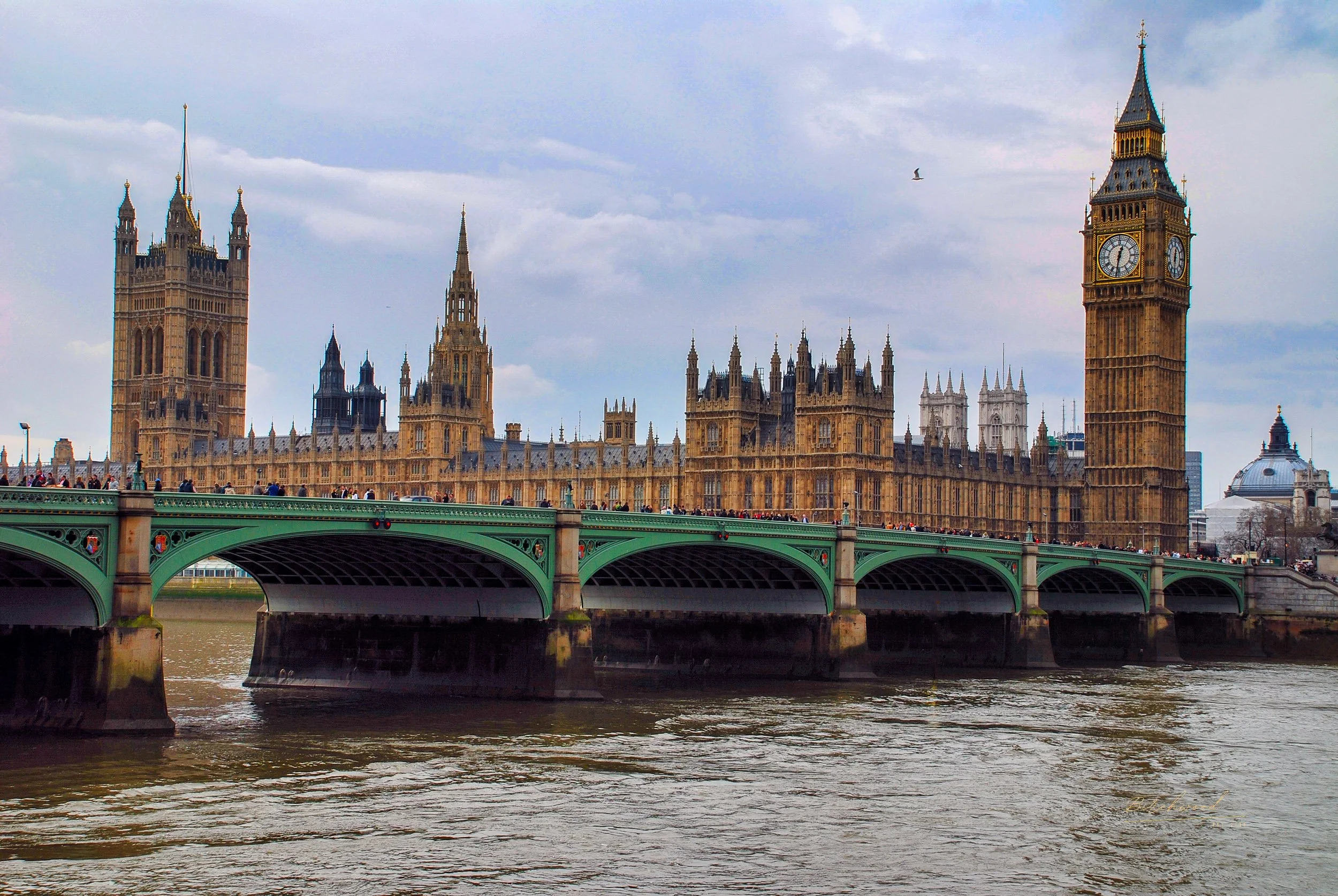 View of the Houses of Parliament and Big Ben in London, England, with a bridge crossing the river in the foreground and cloudy skies overhead.