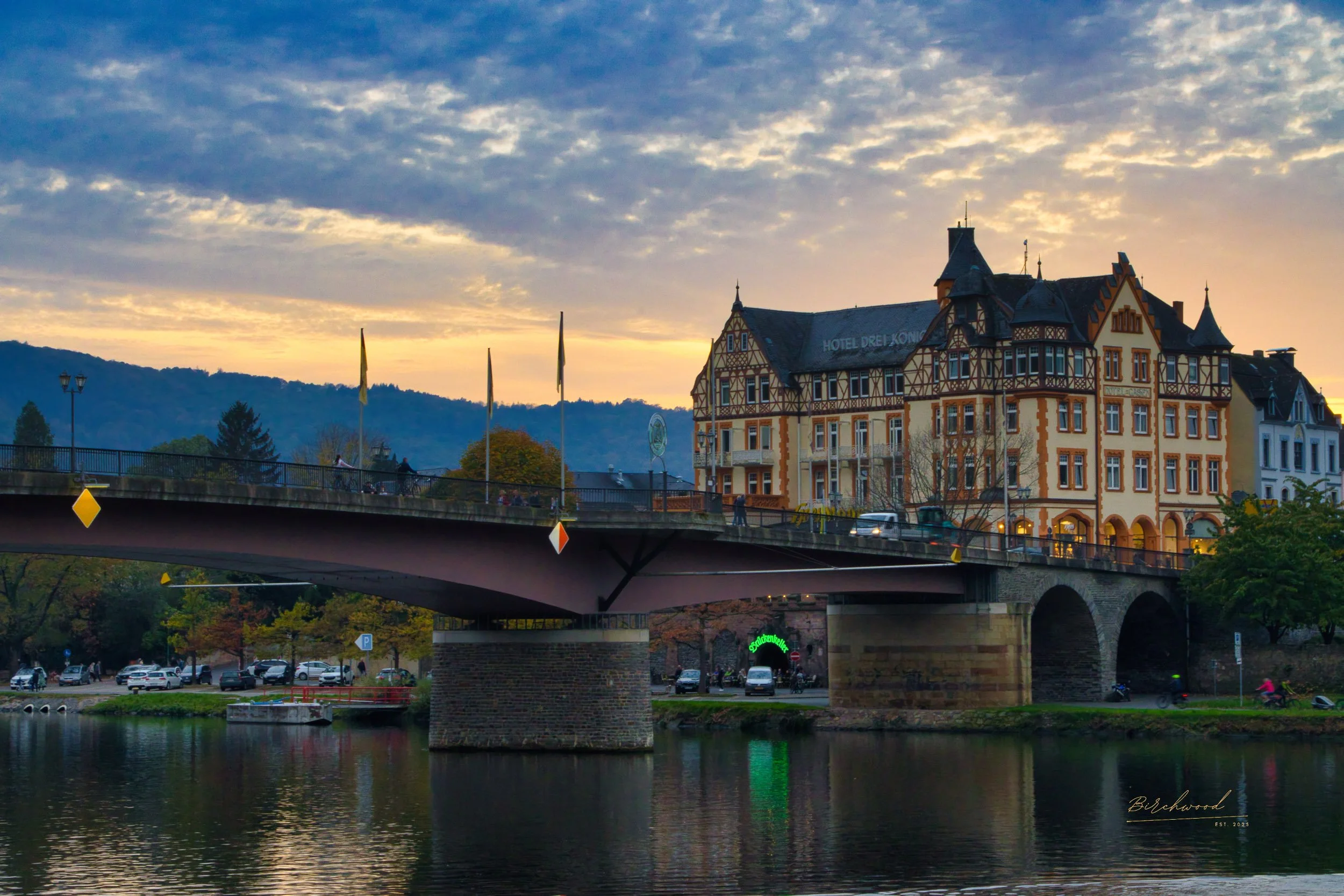 A scenic view Bernkastel-Kues bridge over the Moselle River with a large, historically styled hotel building in the background during sunset, with mountains in the distance.