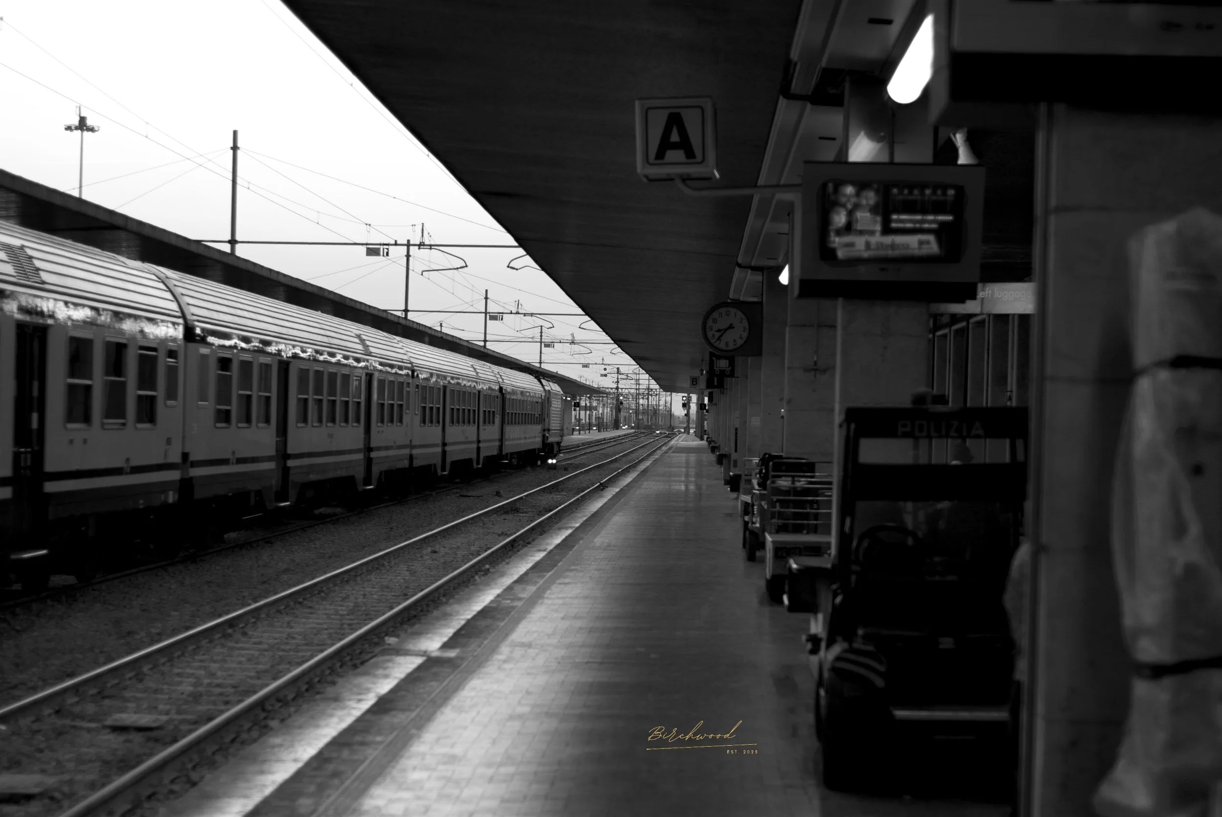 A black and white photo of the Venice, Italy train station platform with a train on the left and a shelter with luggage and a police cart on the right. Overhead sign displays 'A' and a clock shows 4:45.
