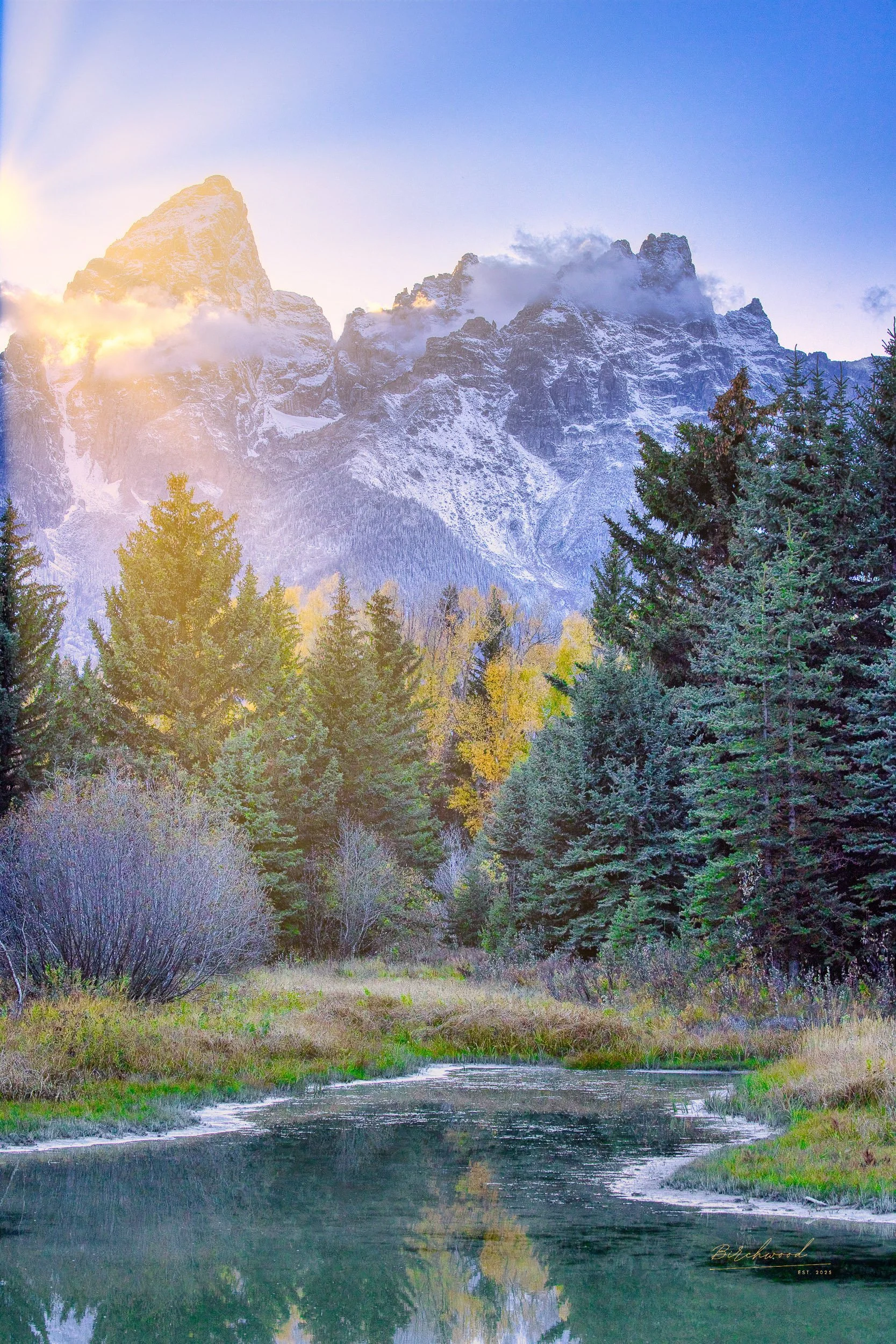 Snow-capped mountains of Grand Teton National Park with clouds at sunset, lush green trees and fall colors surrounding a calm pond.