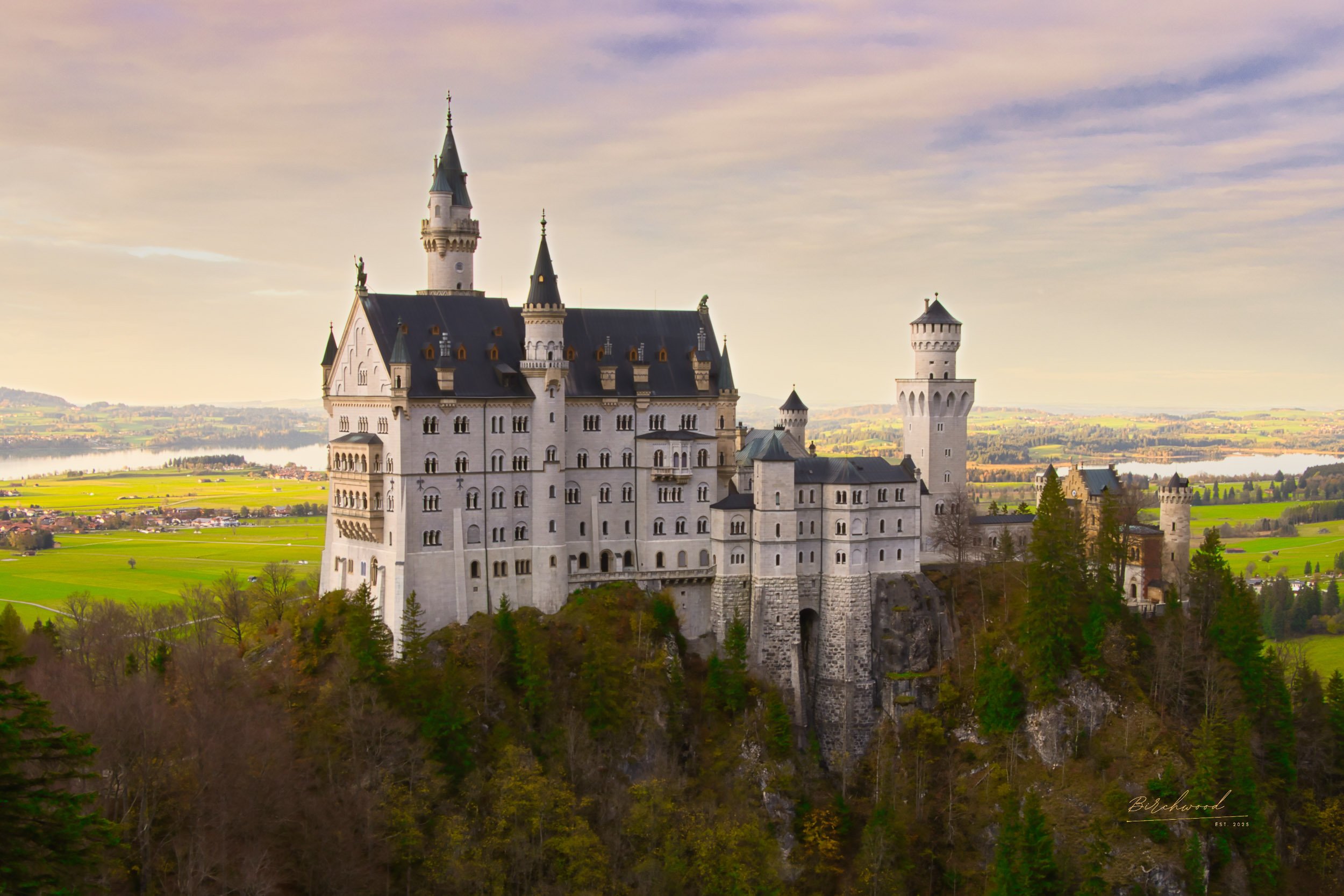 Neuschwanstein castle in Germany with multiple towers and turrets, situated on a rocky hill surrounded by trees, with a landscape of fields and a river in the background during sunset.