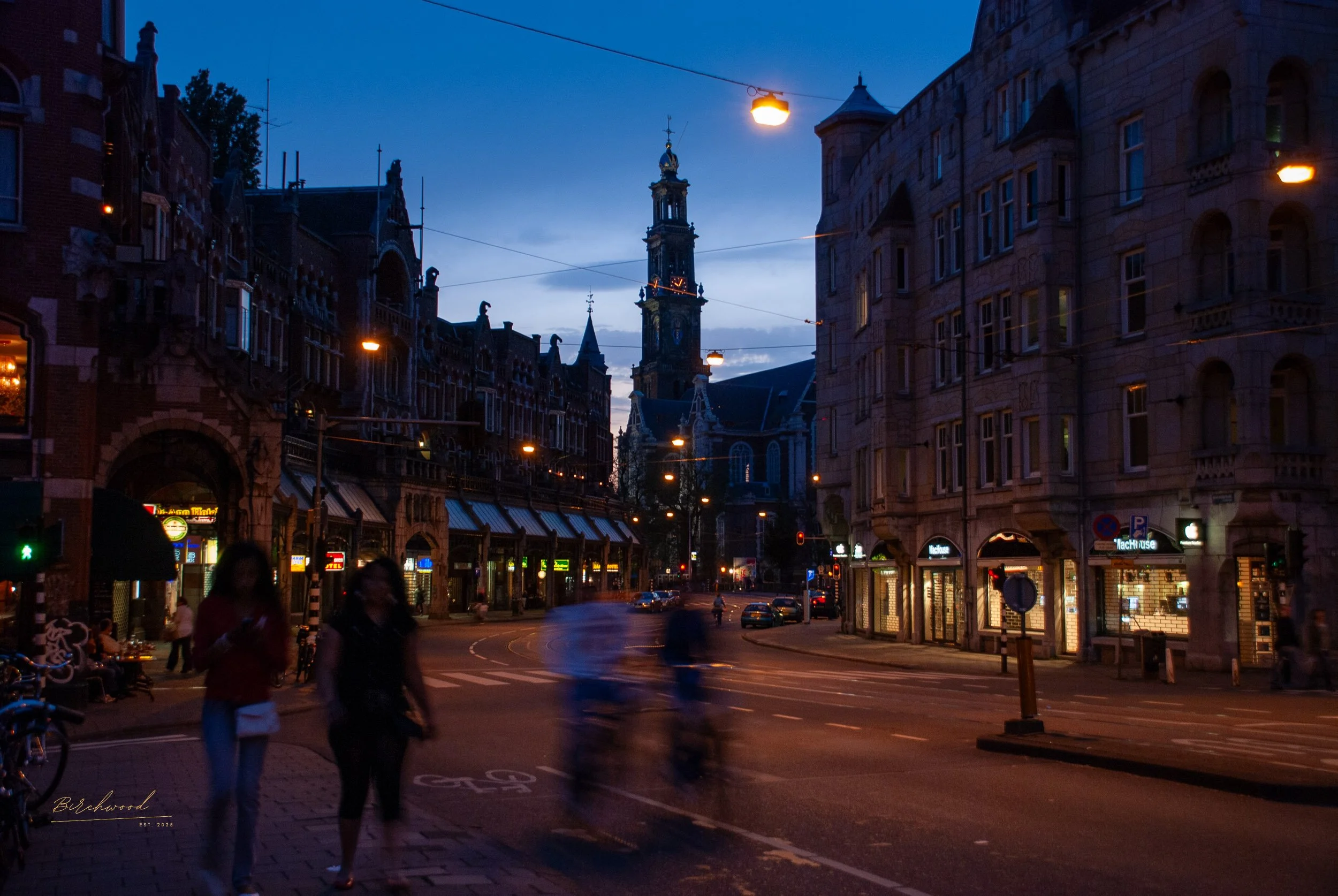 A typical Amsterdam street scene during dusk with blurred pedestrians, parked bicycles, and historic buildings illuminated by streetlights. A tall clock tower is visible in the background.