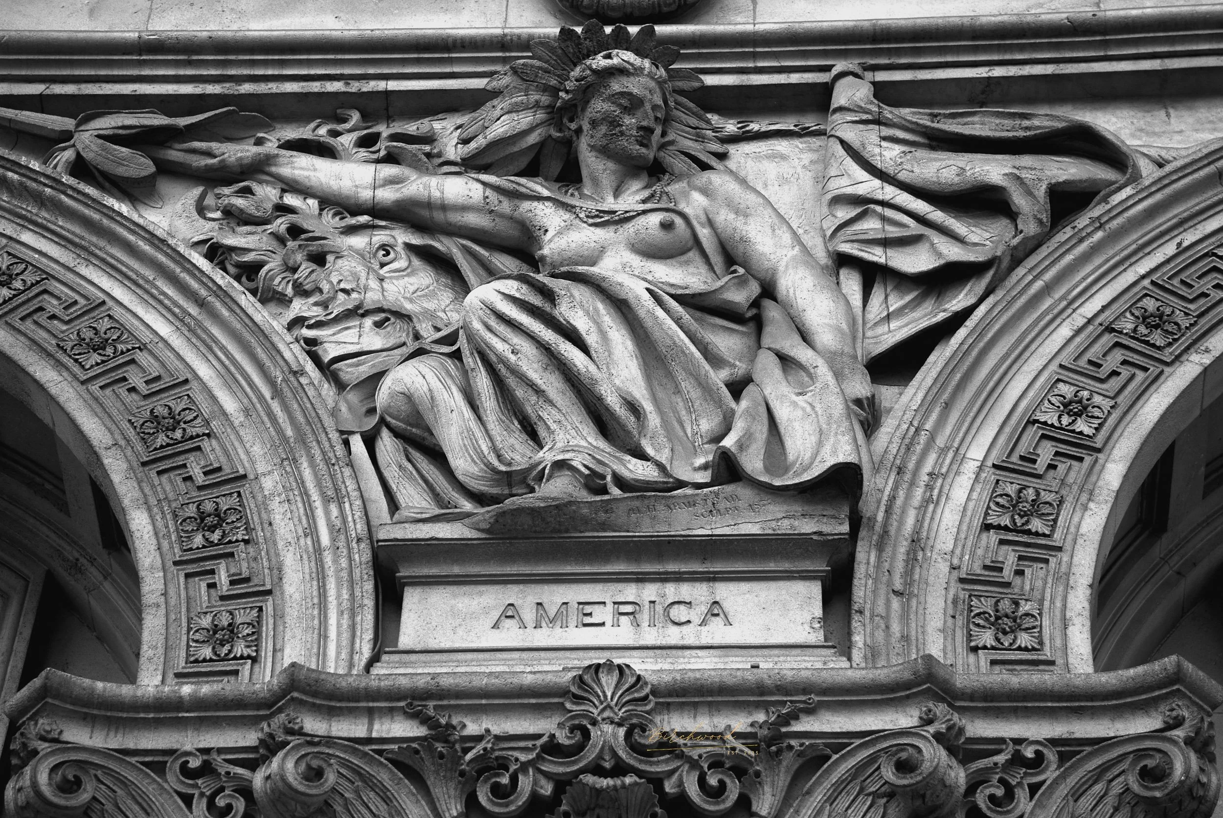 Photograph of an ornate architectural sculpture in London depicting a woman with flowing hair, a crown of feathers, and a draped garment. The woman is seated with one arm extended and the word 'AMERICA' inscribed on the base of the sculpture.