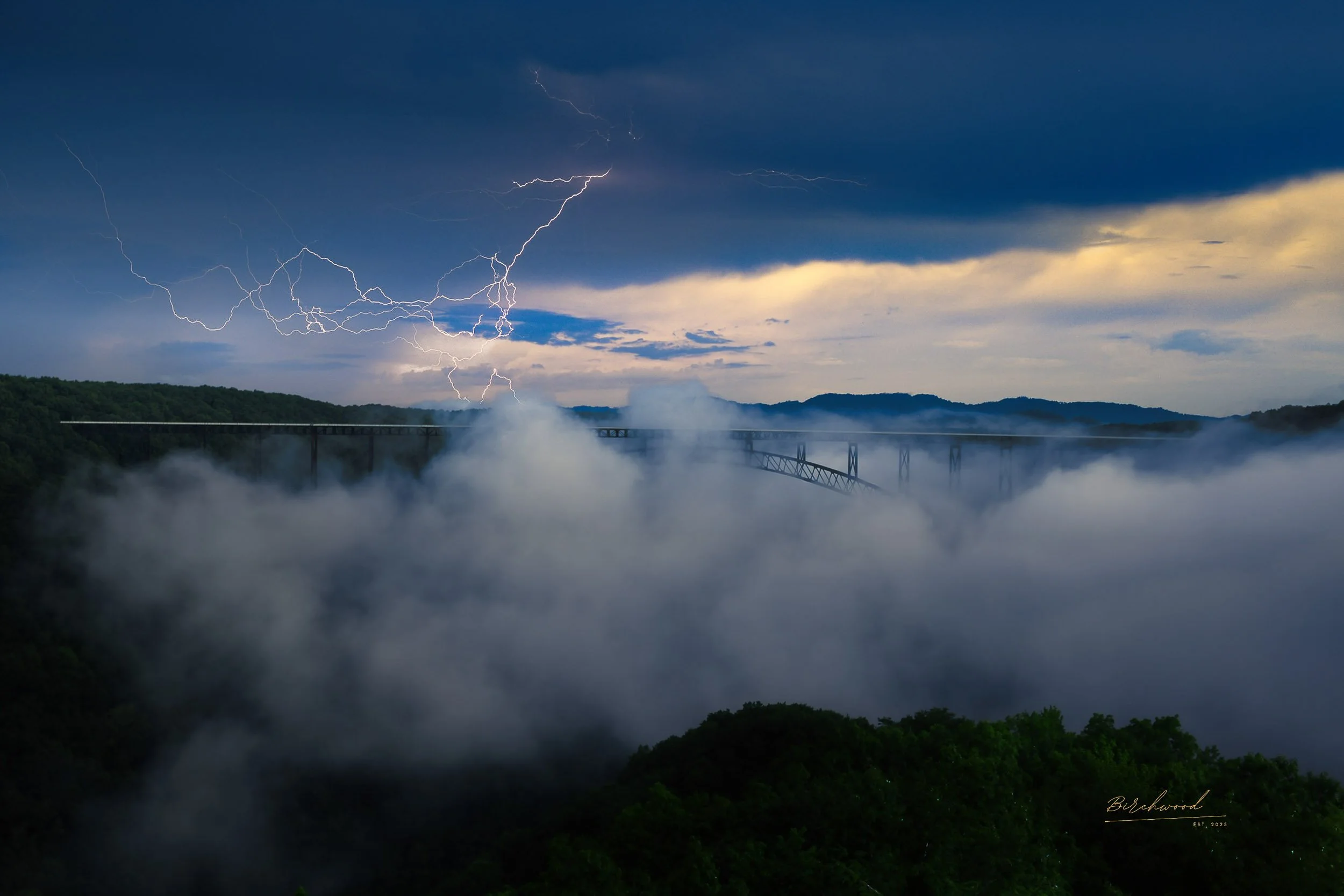 Lightning strikes over a foggy river valley with the New River Gorge National Park bridge and hills in the background during a stormy sky.