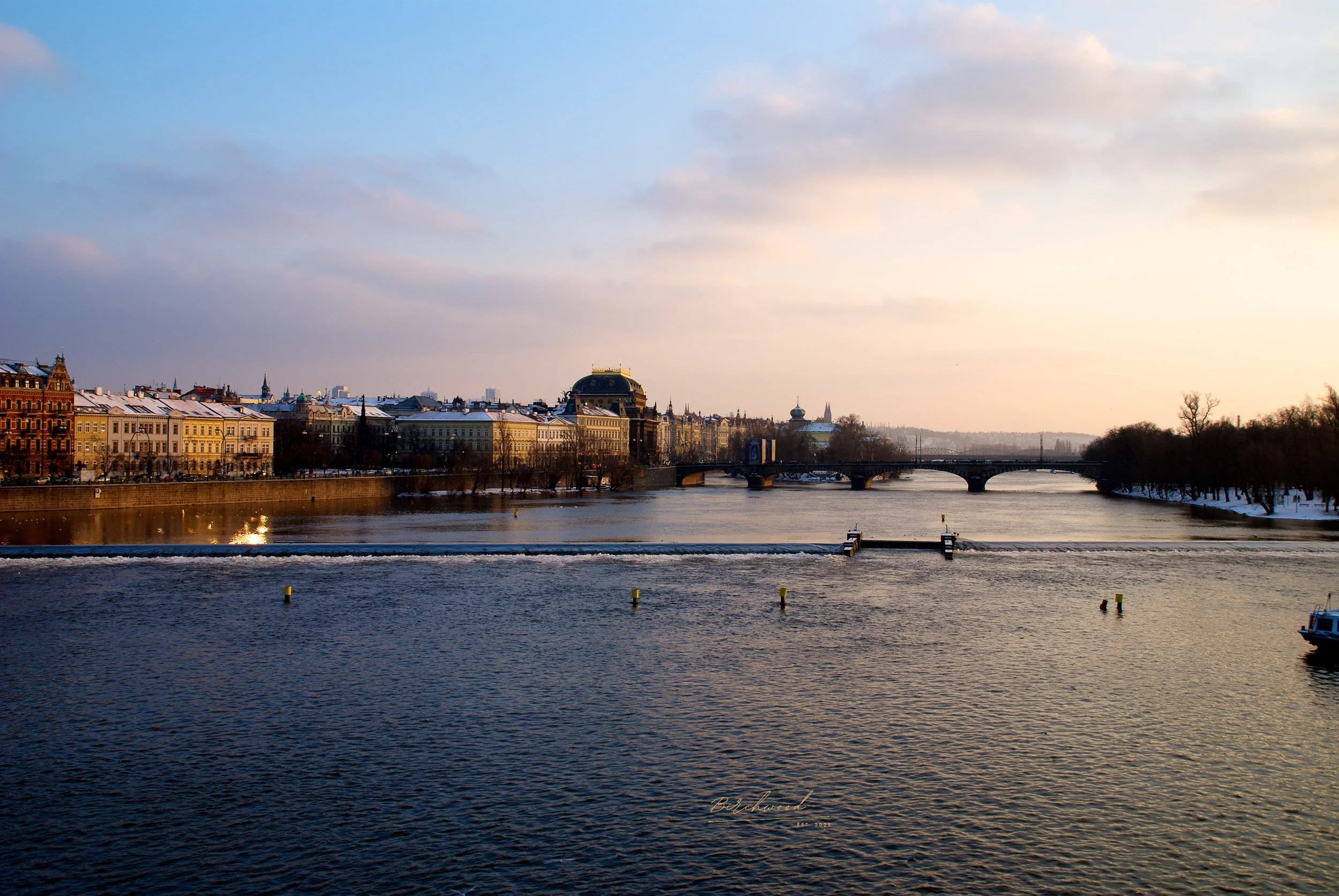 A cityscape of The Vltava river in Prague with historic buildings along the riverbank, and a bridge over the river under a partly cloudy sky.