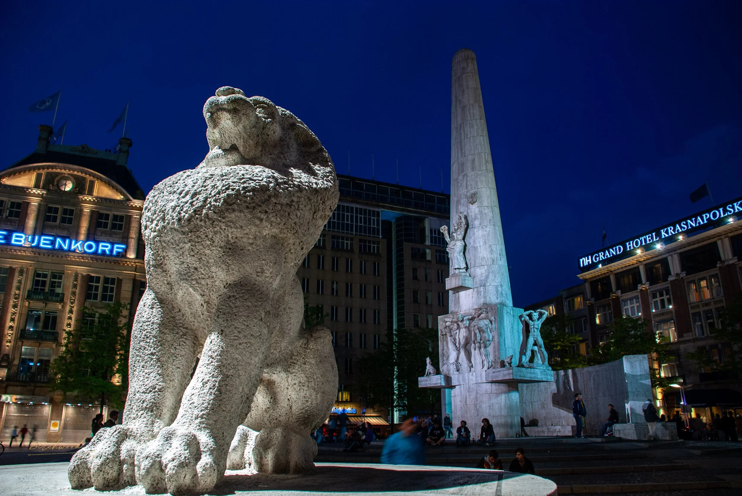 Night view of a city square in Amsterdam with a large stone lion statue in the foreground, and a tall obelisk monument with statues on it in the background. Buildings with illuminated signs, including 'NH Grand Hotel Krasnapolski' and 'Bjenkork,'.